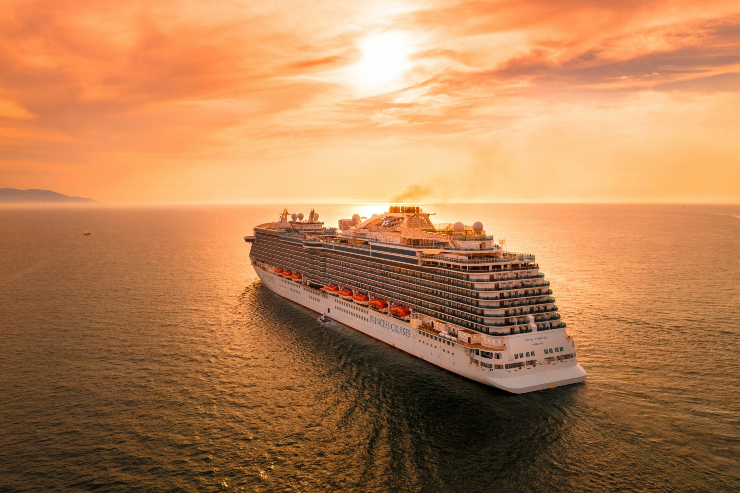A large white cruise ship sails on calm ocean waters at sunset, with an orange sky and the sun glowing near the horizon.