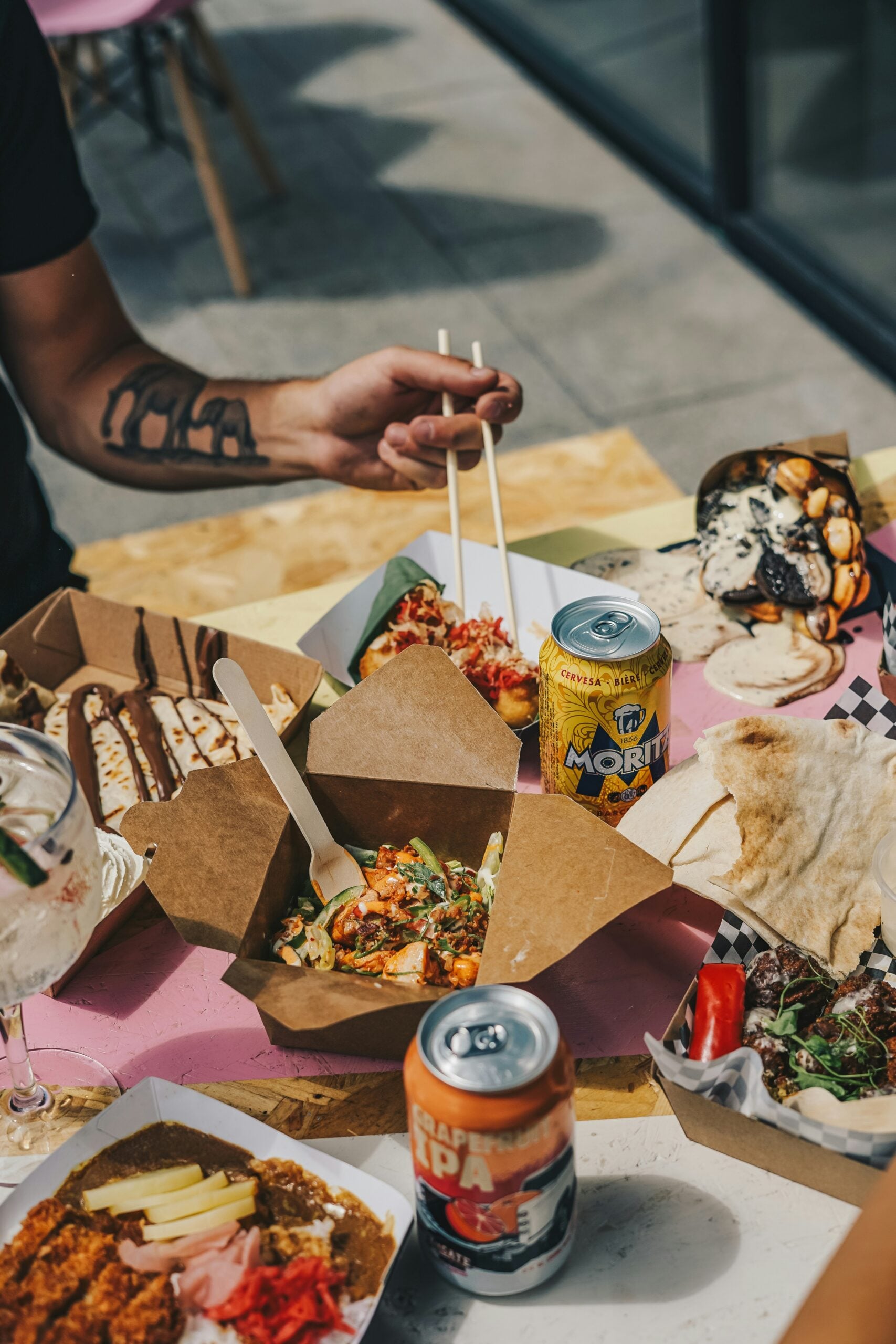 A person with a tattooed arm uses chopsticks to eat from a takeout box. Various street foods, pita bread, and canned drinks are spread on the table. The scene is casual and outdoors.