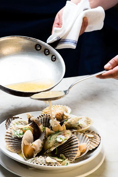 A person pours sauce from a metal pan over a plate of open shellfish on a white marble surface, using a spoon to guide the sauce.