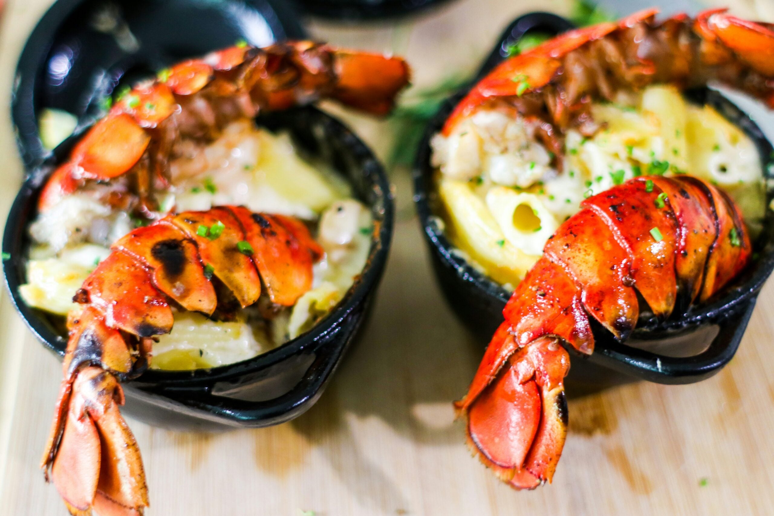 Two small black dishes filled with creamy pasta, each topped with a grilled lobster tail and garnished with chopped chives, set on a light wooden surface.