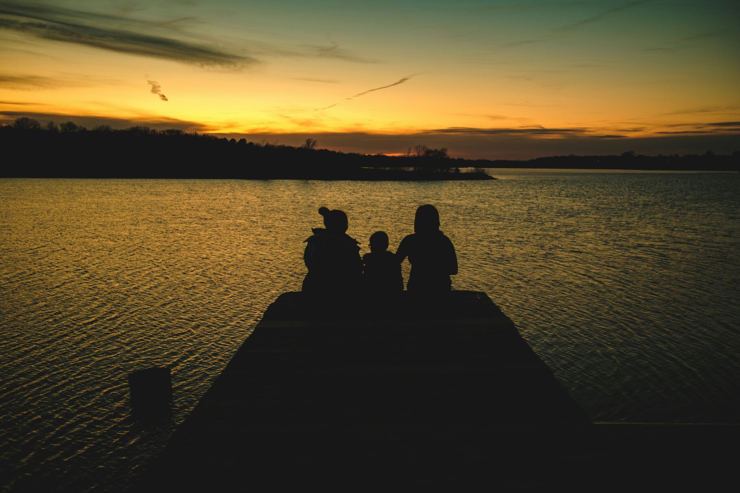Three people sit together at the end of a dock, silhouetted against a golden sunset over a calm lake, with trees visible along the distant shoreline.