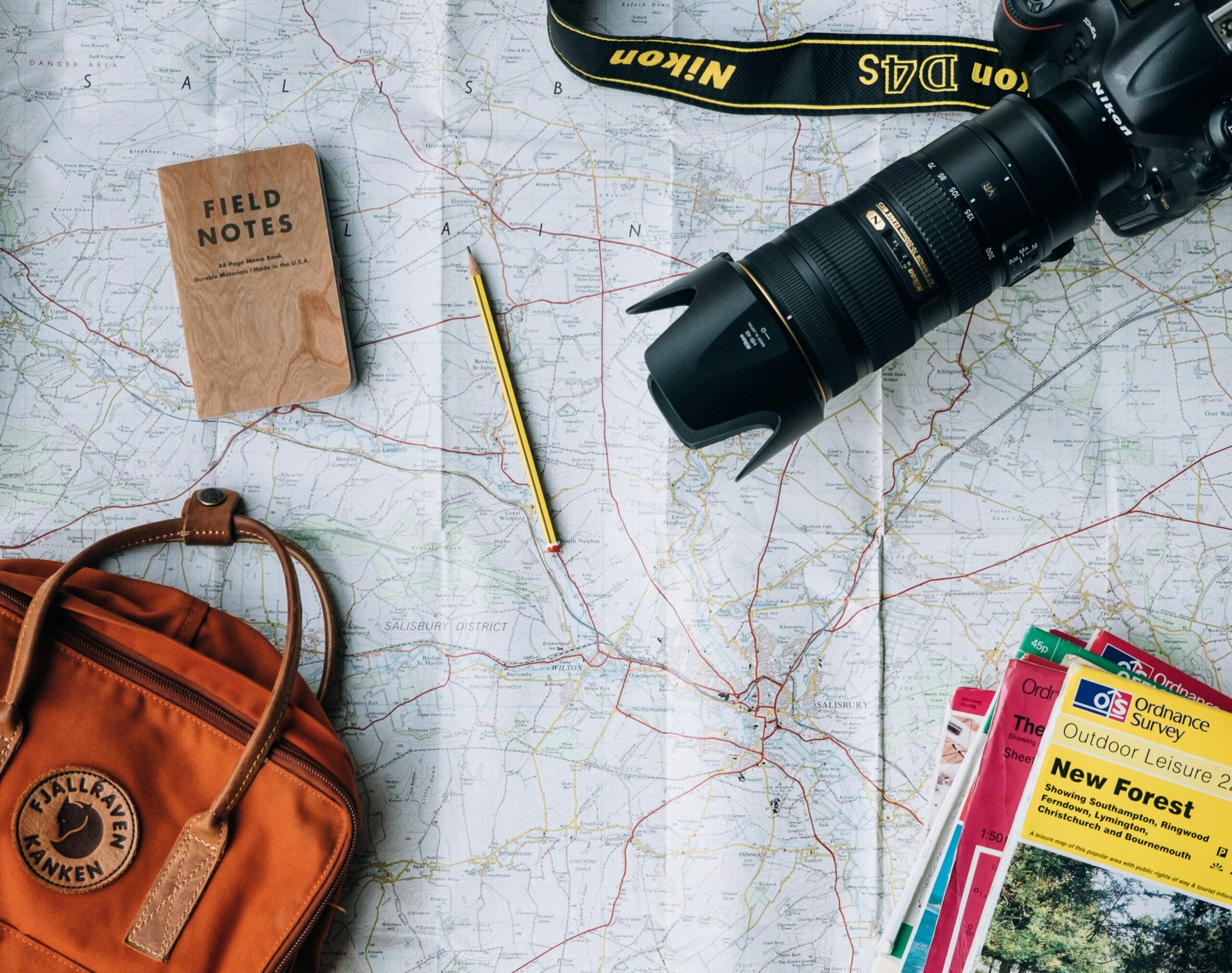 A map spread out on a surface with a camera, a field notes notebook, a pencil, an orange backpack, and several guidebooks arranged on top, suggesting travel or adventure planning.