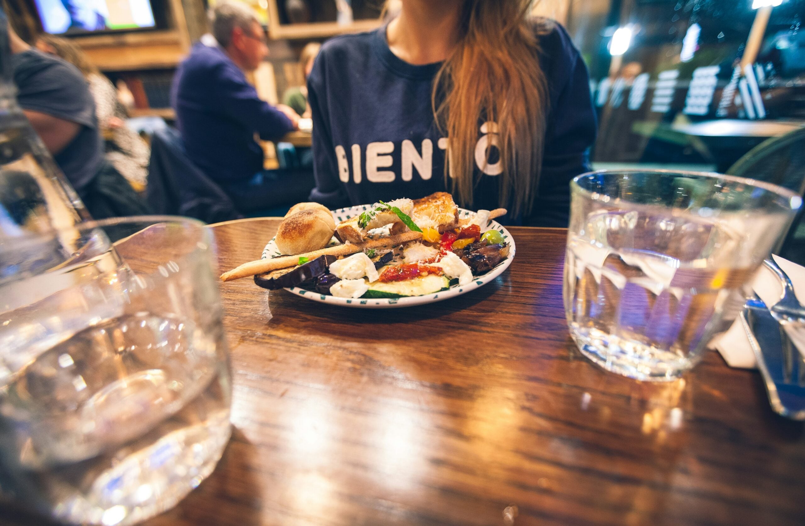 A woman sits at a wooden table in a restaurant with a plate of assorted food in front of her, flanked by two glasses of water. The background shows other diners and a cozy, warmly-lit atmosphere.