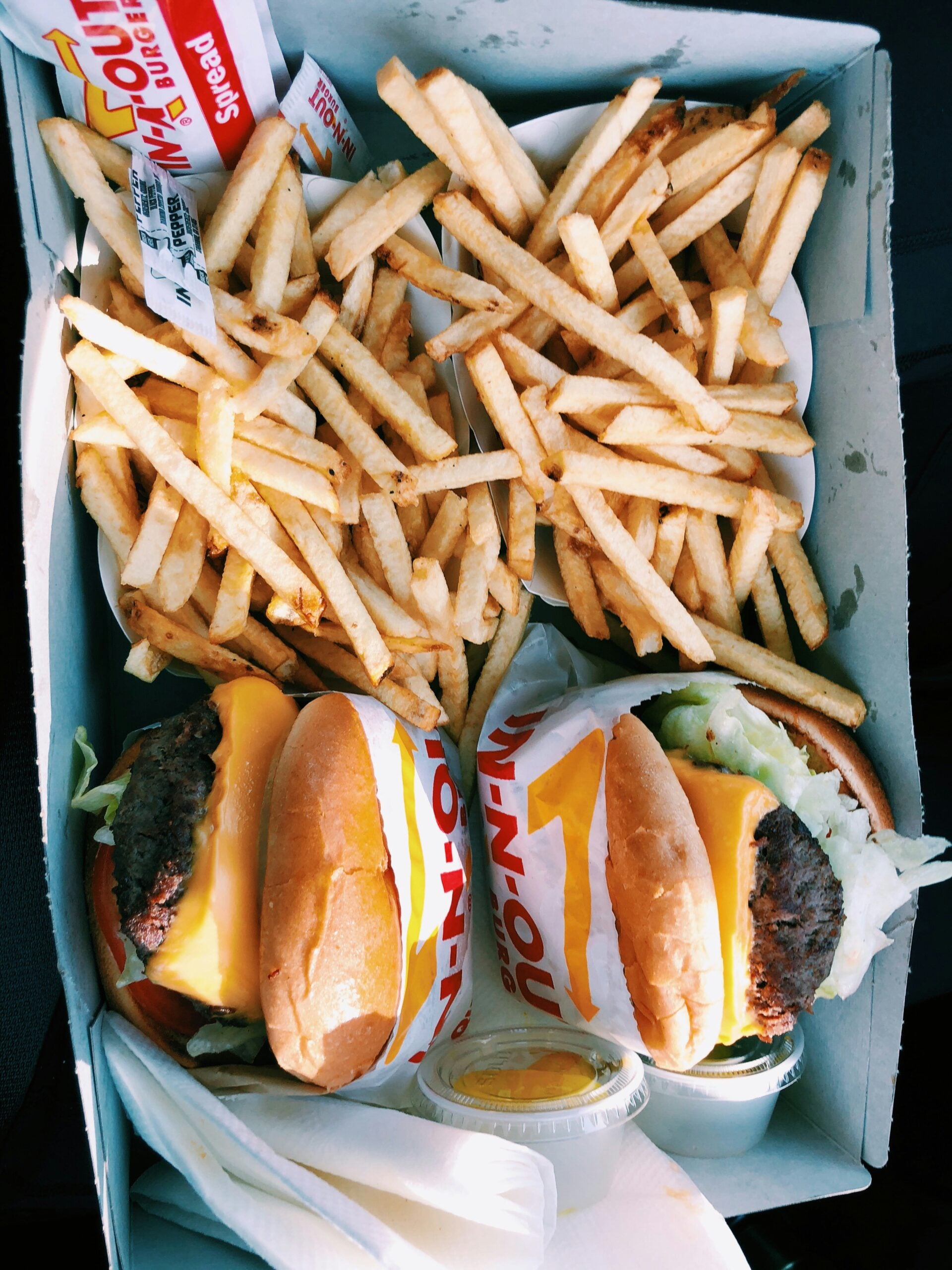 A takeout box from In-N-Out contains two cheeseburgers wrapped in branded paper, two servings of golden French fries, ketchup packets, and small condiment containers.