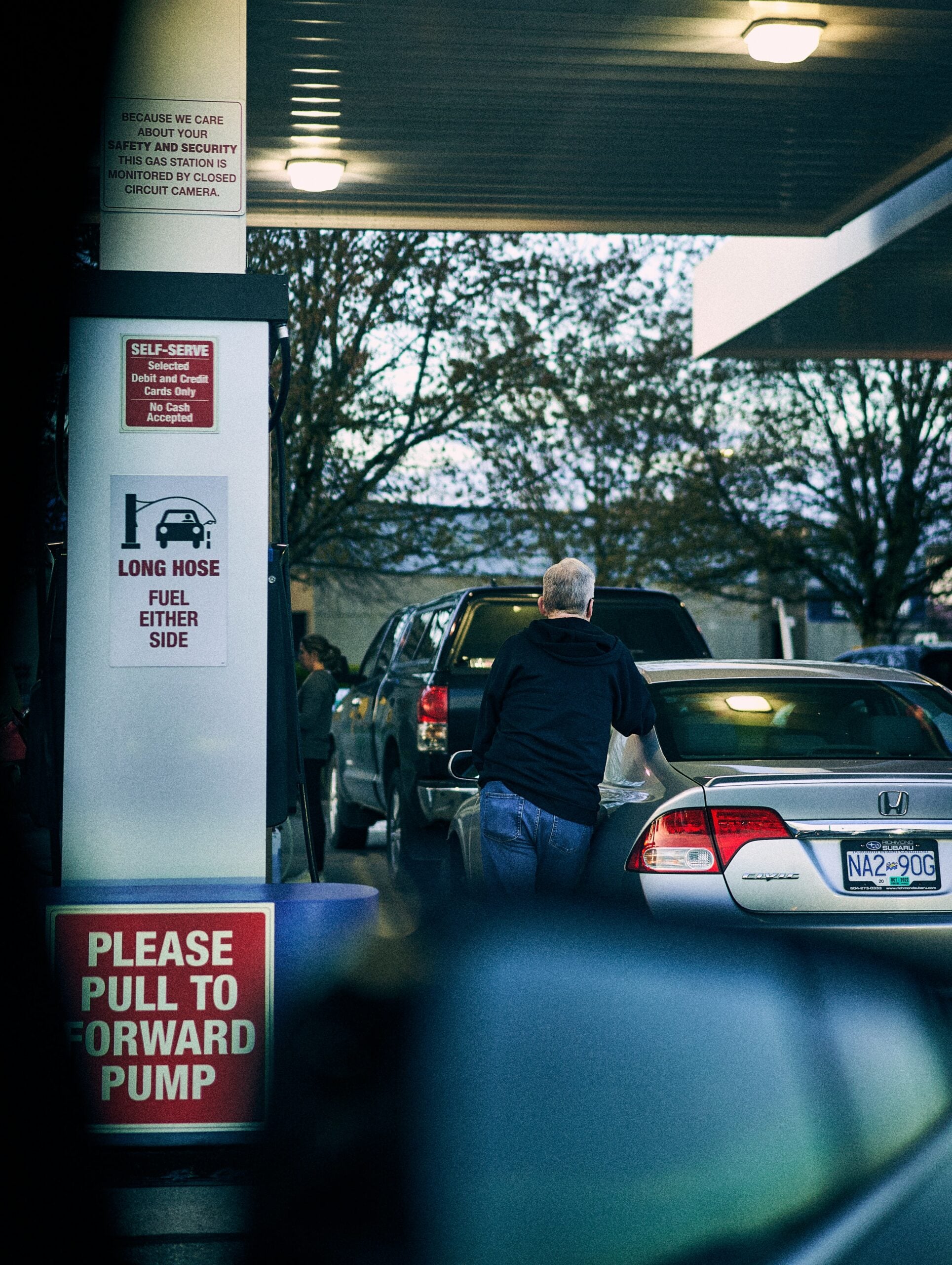 A person refuels a silver car at a gas station pump, with visible signs reading "LONG HOSE FUEL EITHER SIDE" and "PLEASE PULL TO FORWARD PUMP." Trees and parked vehicles are in the background.