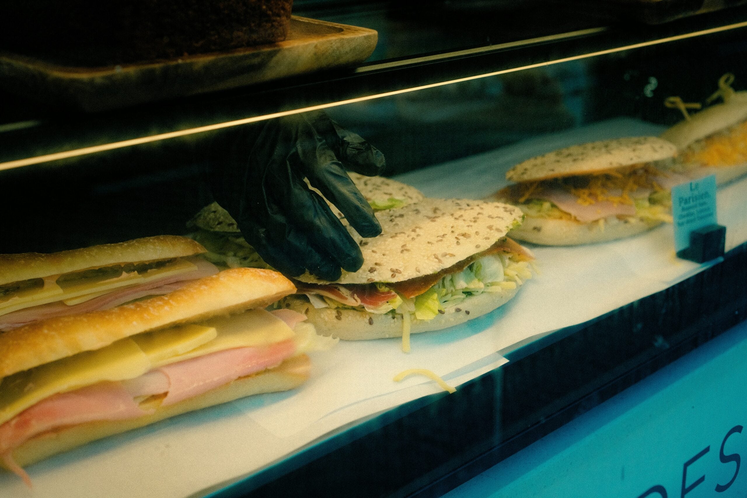 A person wearing a black glove assembles sandwiches with cold cuts, cheese, and lettuce behind a glass display at a deli or sandwich shop.