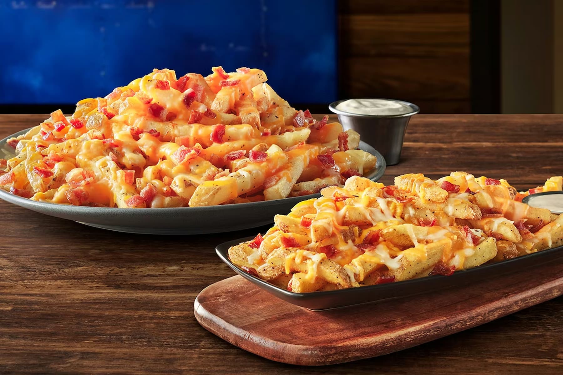 Two plates of loaded fries topped with melted cheese, diced bacon, and seasoning sit on a wooden table. A small cup of ranch dipping sauce is placed behind the plates.