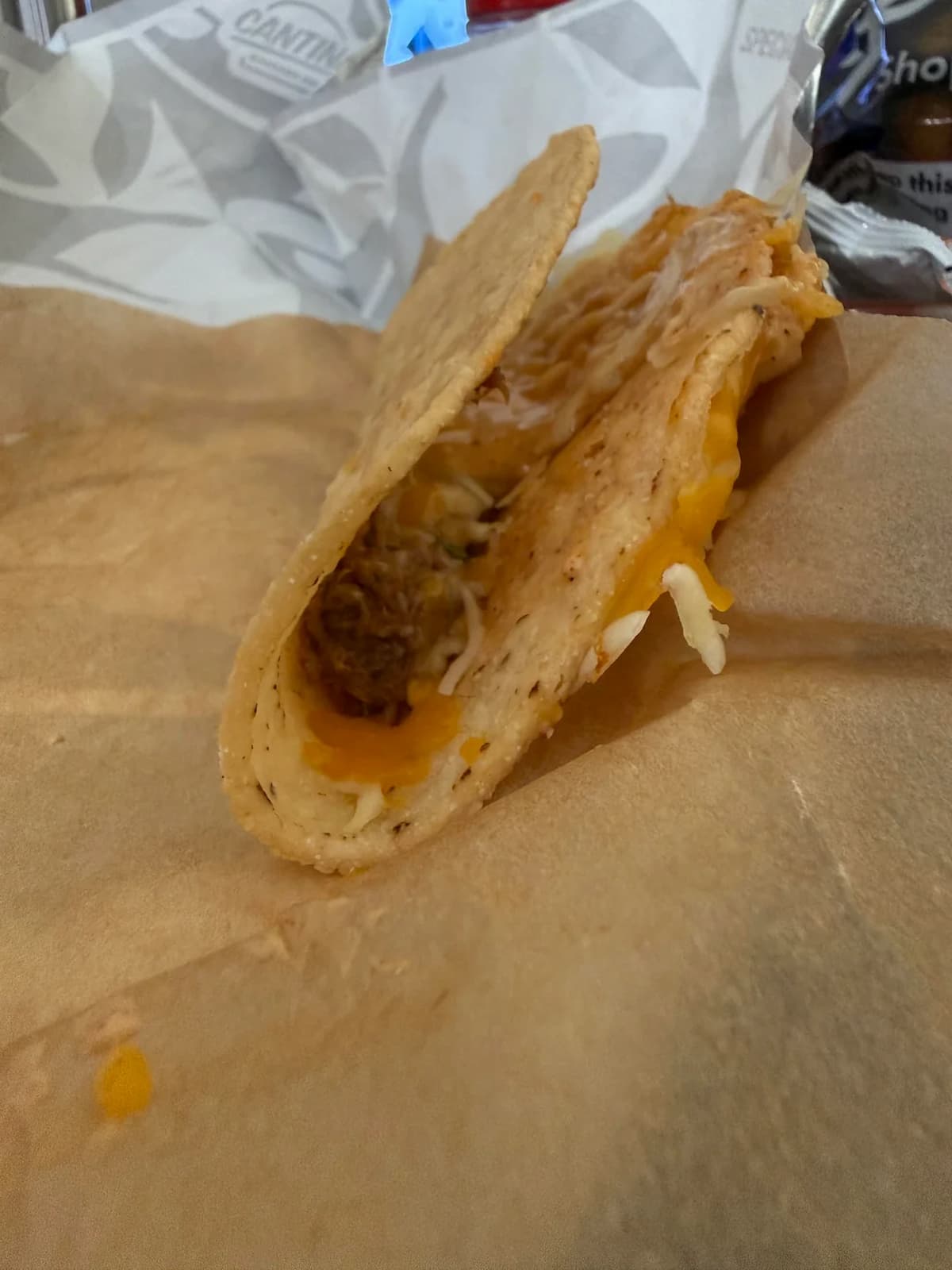 A close-up of a taco with a hard, cheesy shell filled with shredded cheese and meat, placed on brown parchment paper. The background includes a food wrapper and part of a drink.