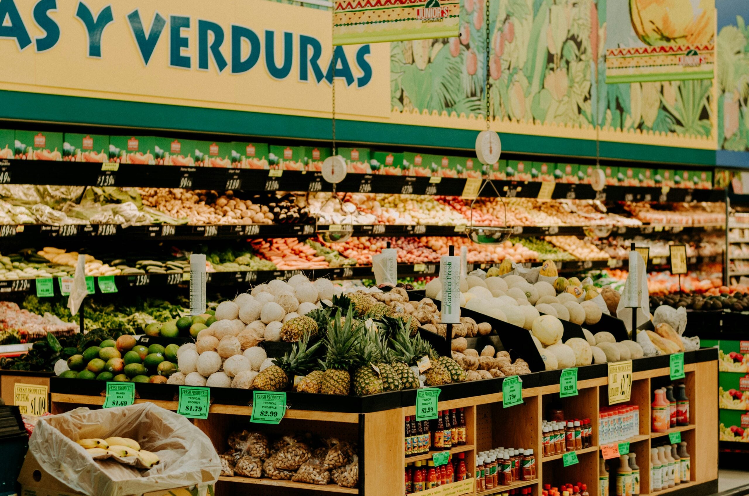 A supermarket produce section displays a variety of fruits and vegetables, including coconuts, pineapples, melons, avocados, and limes, with signs indicating prices.