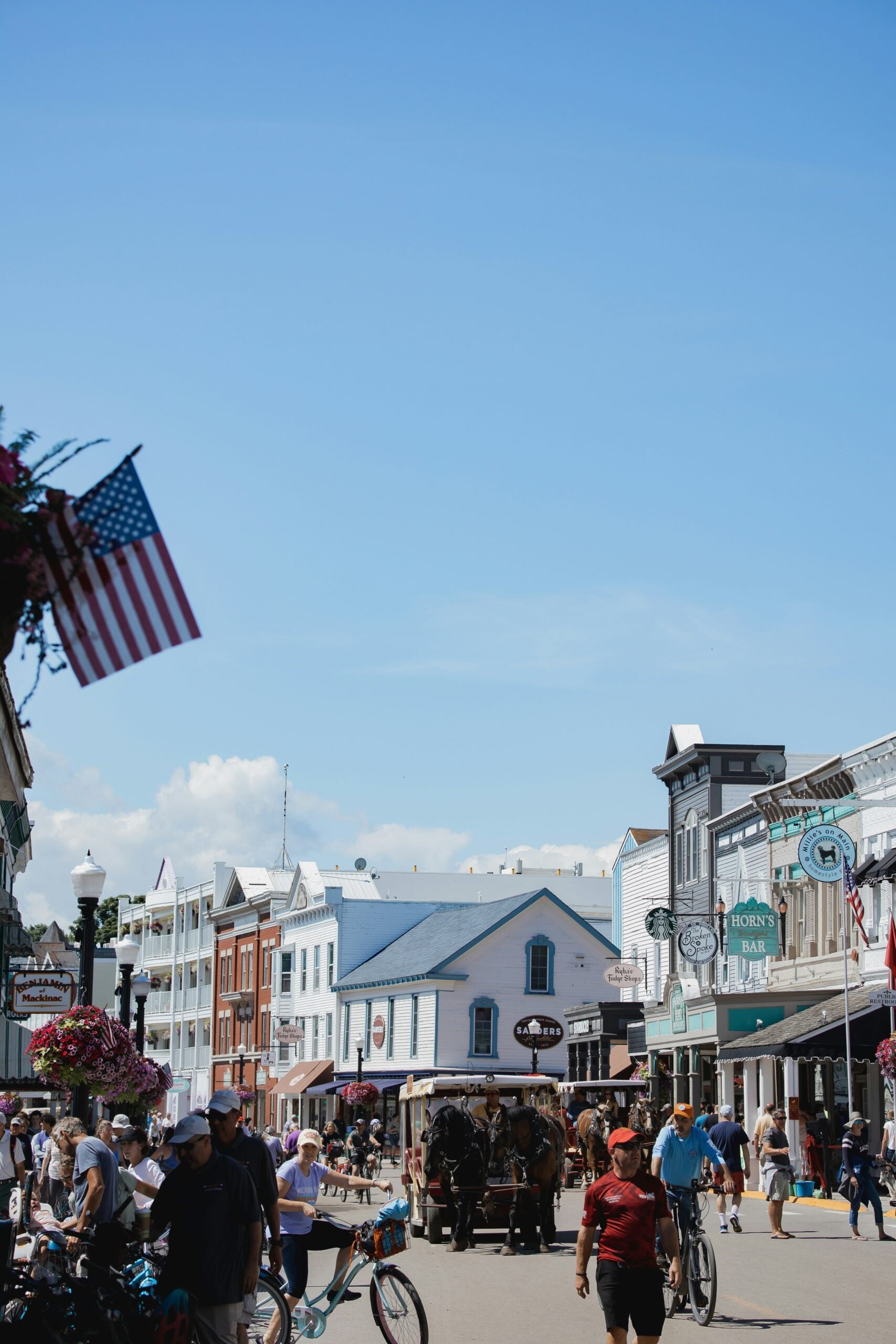 A busy street scene in a historic town with people walking and biking, horse-drawn carriages, old-fashioned buildings, hanging flower baskets, and an American flag on a sunny day.