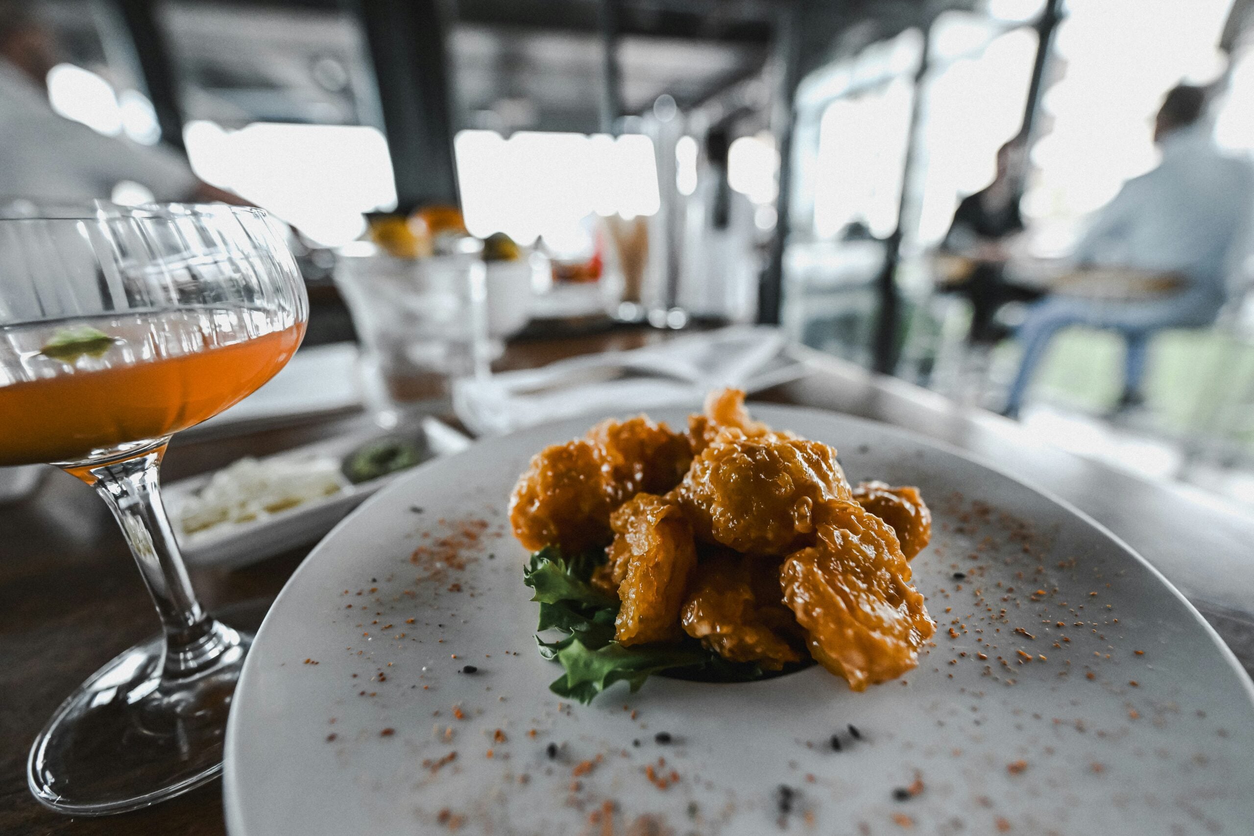 A close-up of a plate of fried shrimp garnished with greens and seasoning, with a cocktail glass on the side. The background shows a blurred restaurant setting with people dining.