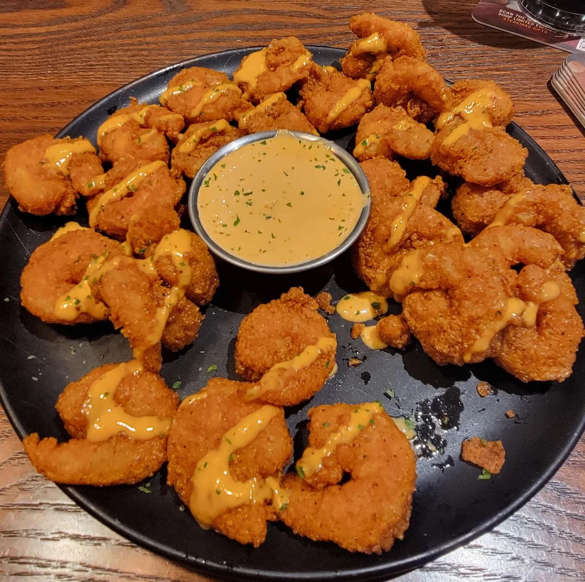 A black plate filled with breaded, fried shrimp drizzled with sauce and garnished with herbs, surrounding a small bowl of dipping sauce, all set on a wooden table.