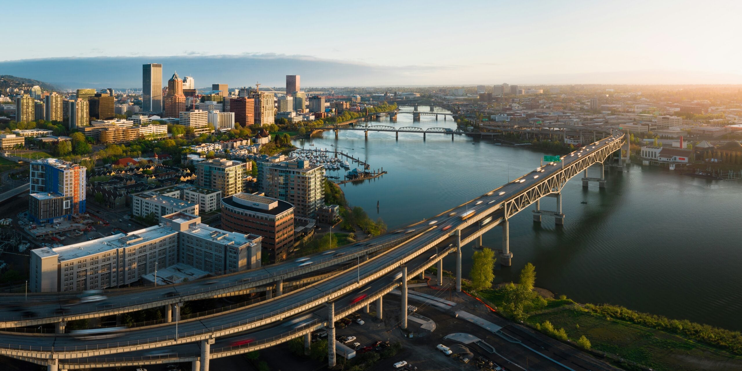 Aerial view of a city with tall buildings, highways, and bridges over a wide river during sunrise or sunset, with sunlight illuminating the skyline and water.