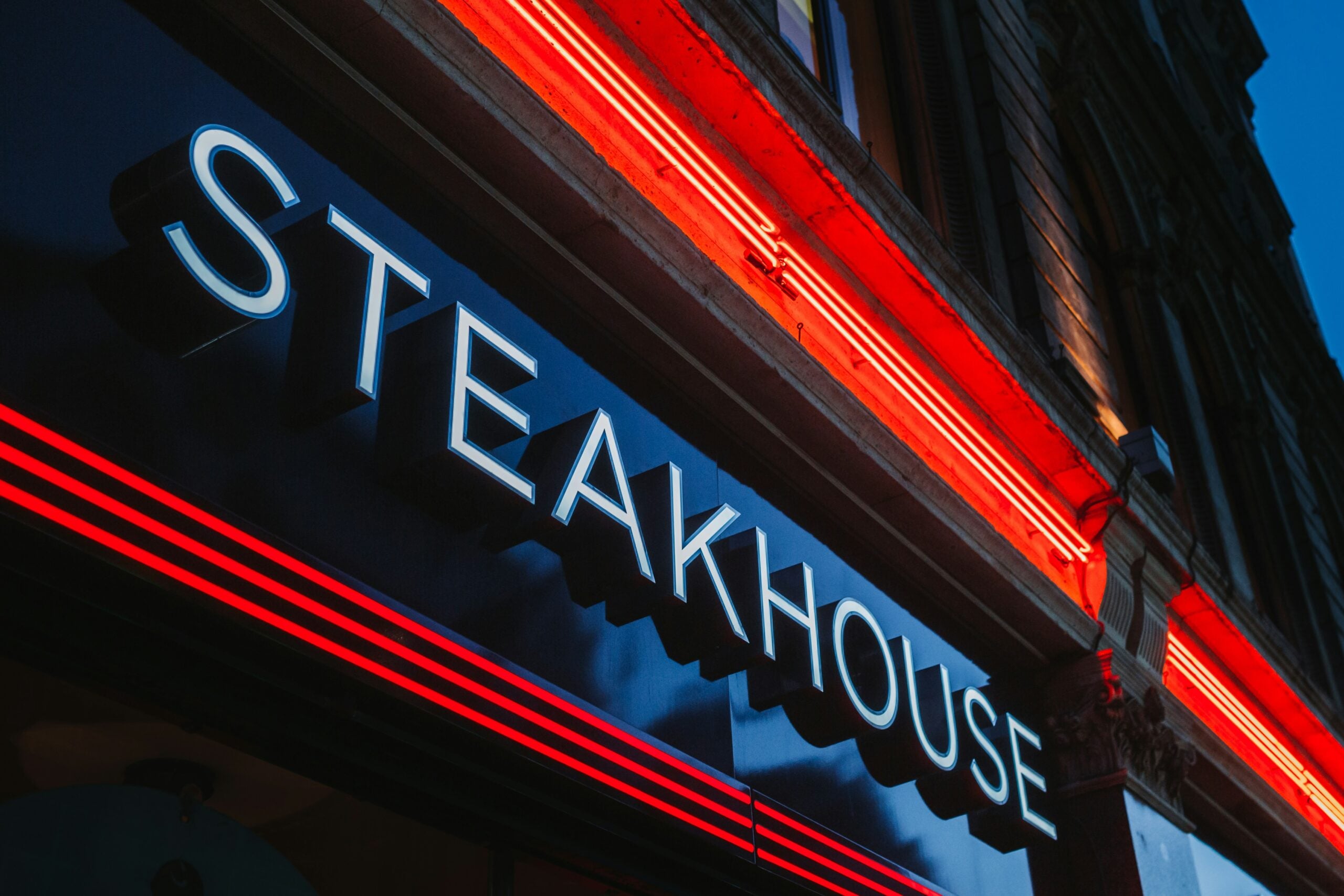 A close-up of a modern steakhouse sign, illuminated with bright white and red neon lights, mounted on the exterior of a building at dusk.