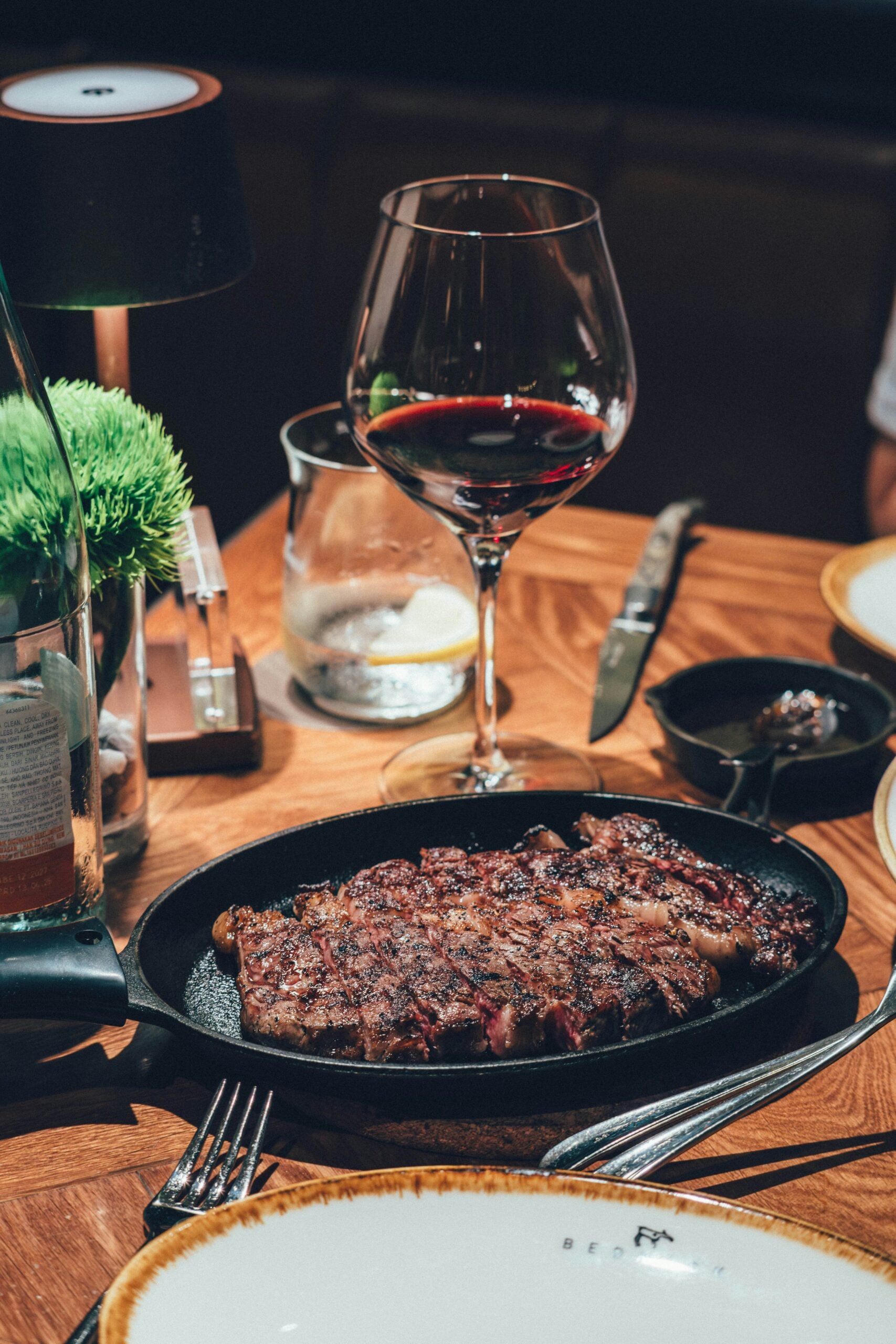 A grilled steak served on a hot plate sits on a wooden table next to a glass of red wine, a water glass with ice, and a small lamp, creating an elegant dining setting.