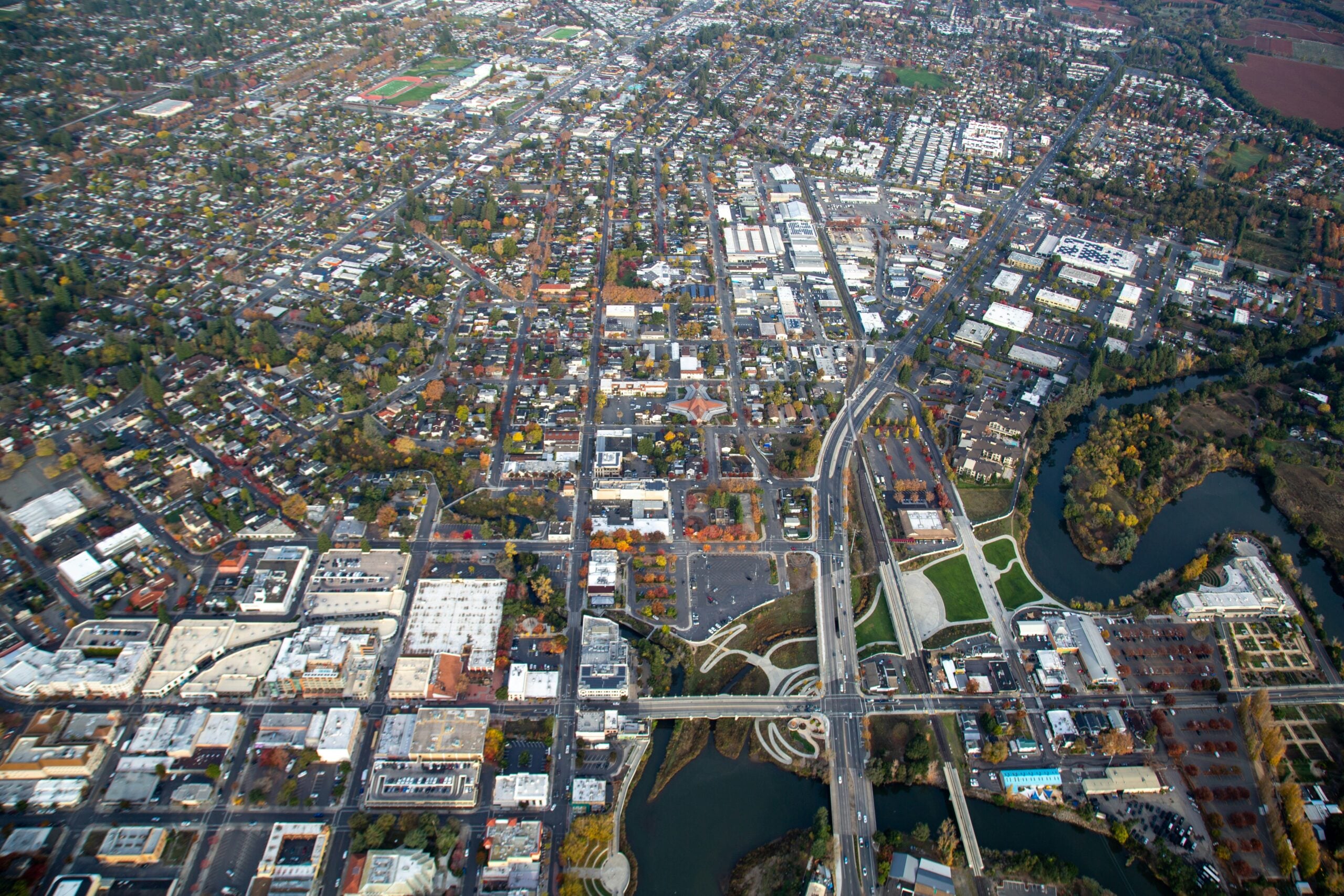 Aerial view of a city with a grid of streets, residential and commercial buildings, a river with bridges, parks, and patches of greenery scattered throughout the urban landscape.
