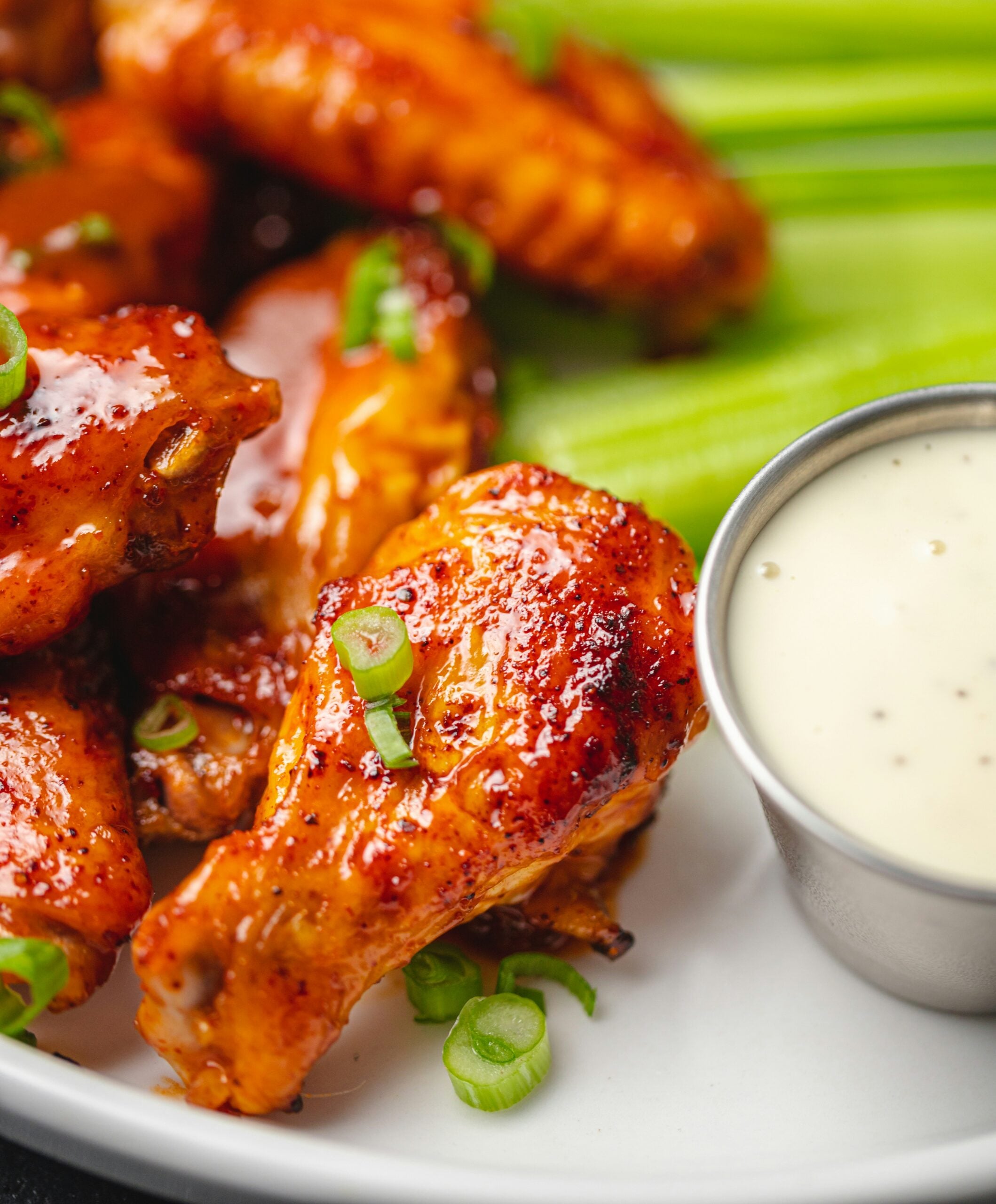 A plate of glazed buffalo chicken wings garnished with sliced green onions, served with fresh celery sticks and a cup of creamy ranch dipping sauce.