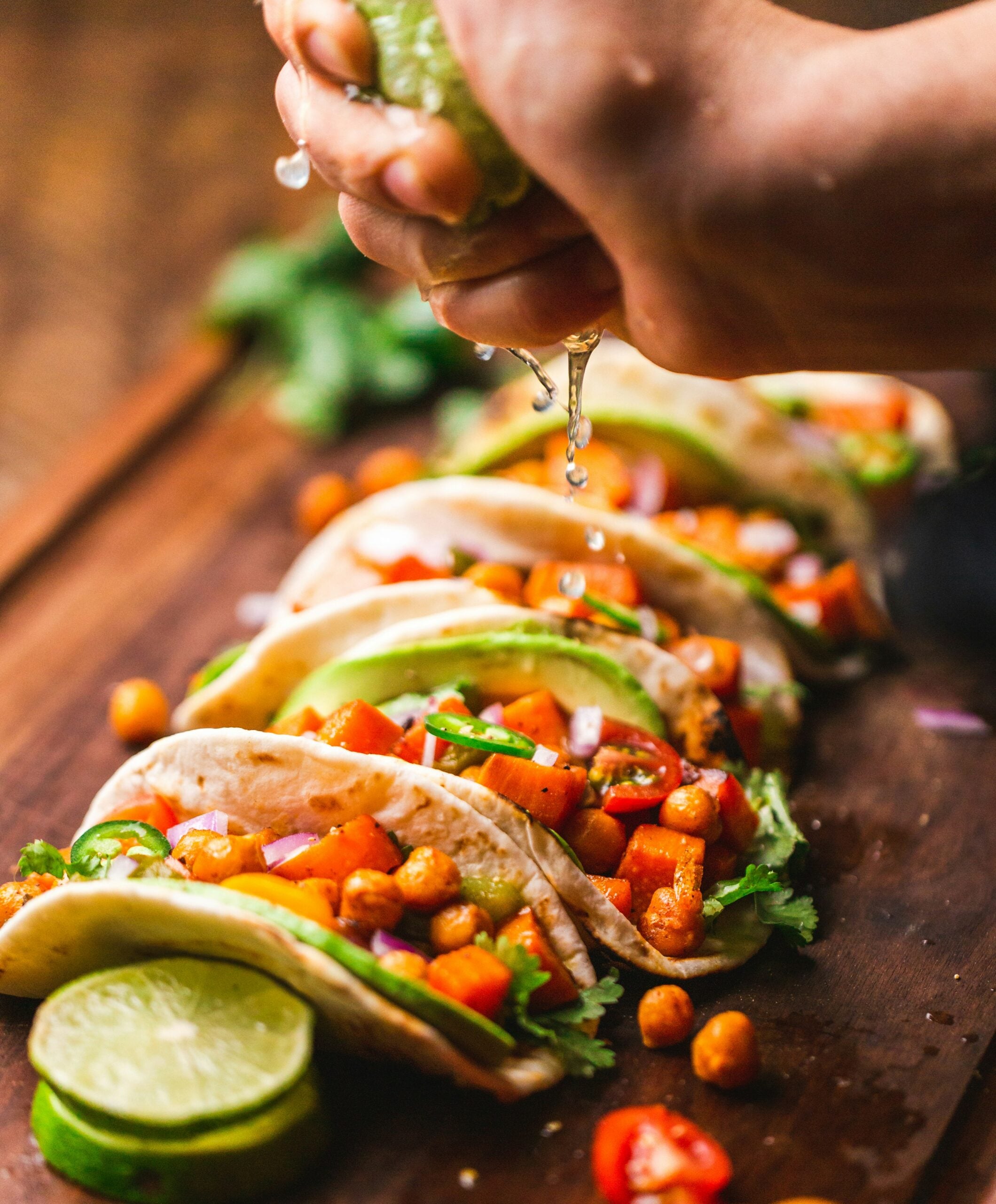 A hand squeezes lime juice over a row of tacos filled with chickpeas, diced vegetables, red onion, cilantro, and sliced jalapeños on a wooden board with lime slices nearby.