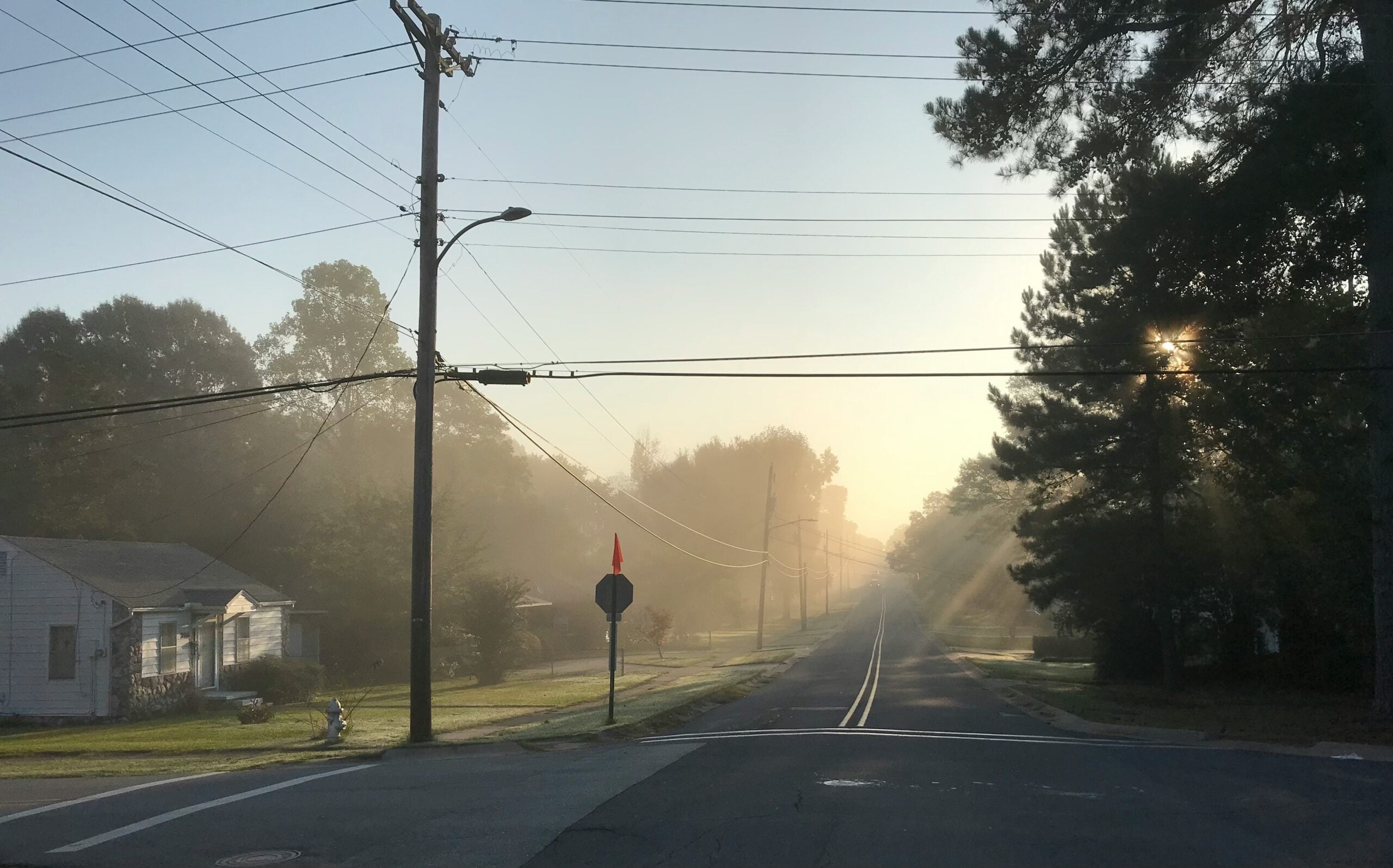 A quiet, empty street at sunrise with mist in the air, power lines crossing overhead, sun rays streaming through tall trees, and small houses along the sidewalk.