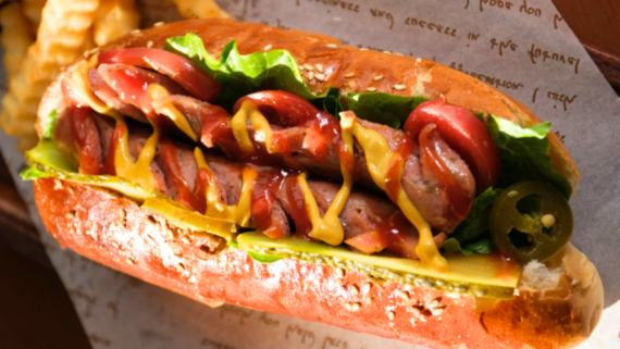 A close-up of a loaded hot dog in a bun, topped with tomato slices, lettuce, mustard, ketchup, pickles, and jalape&ntilde;o slices. Crinkle-cut fries are visible in the background&mdash;a classic bite in one of America&rsquo;s best cheap food cities.