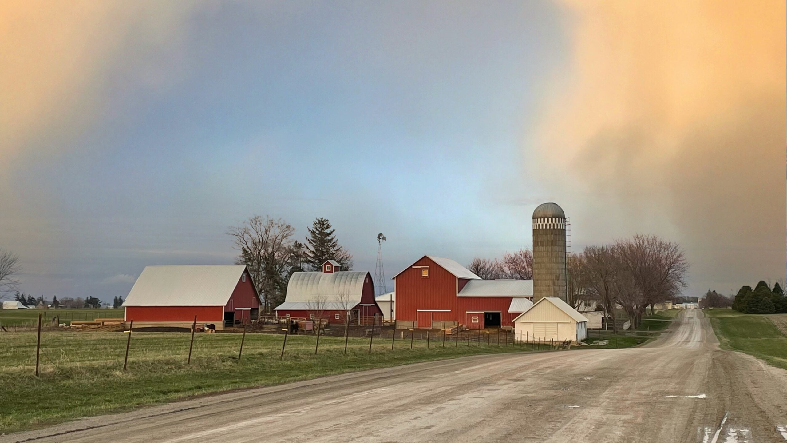 A rural scene with red barns, a silo, and several farm buildings alongside a gravel road, under a sky with soft orange and blue hues. The landscape is green and open, with trees in the background.