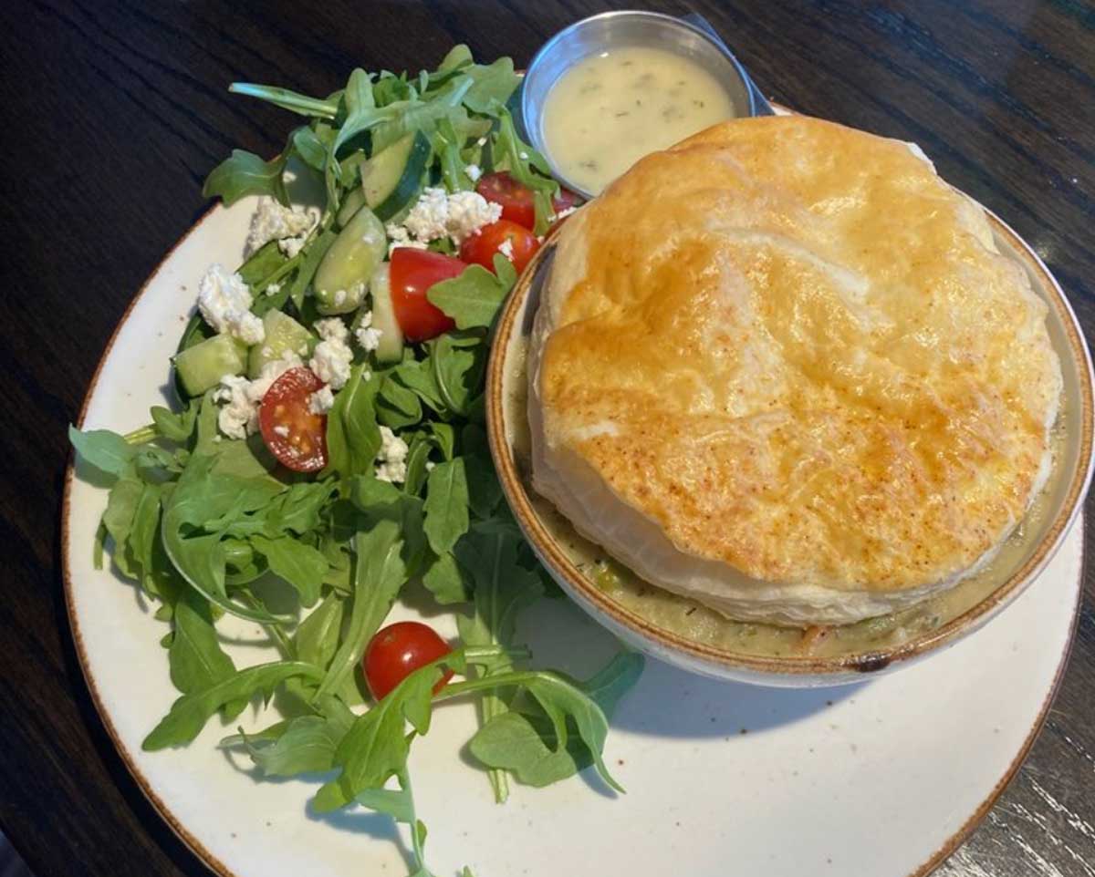 A plate with a golden-brown pot pie in a bowl—just like the best chicken pot pie in every state—served alongside a fresh mixed salad with arugula, cucumber, cherry tomatoes, feta cheese, and a small cup of dressing.