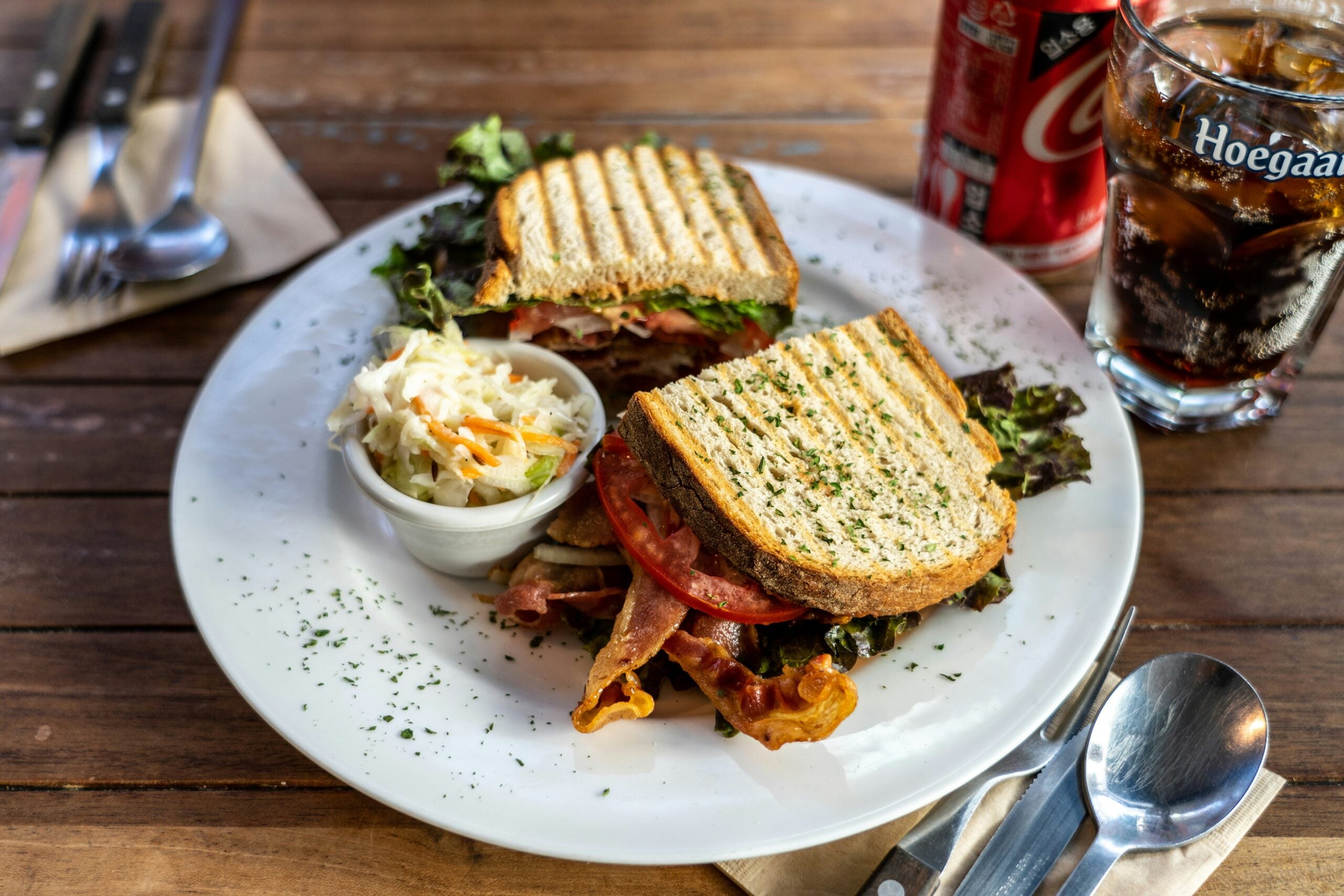 A plate with a toasted sandwich filled with bacon, lettuce, and tomato, served with a side of coleslaw. A glass of cola, a can of Coca-Cola, and cutlery are on the wooden table beside the plate.