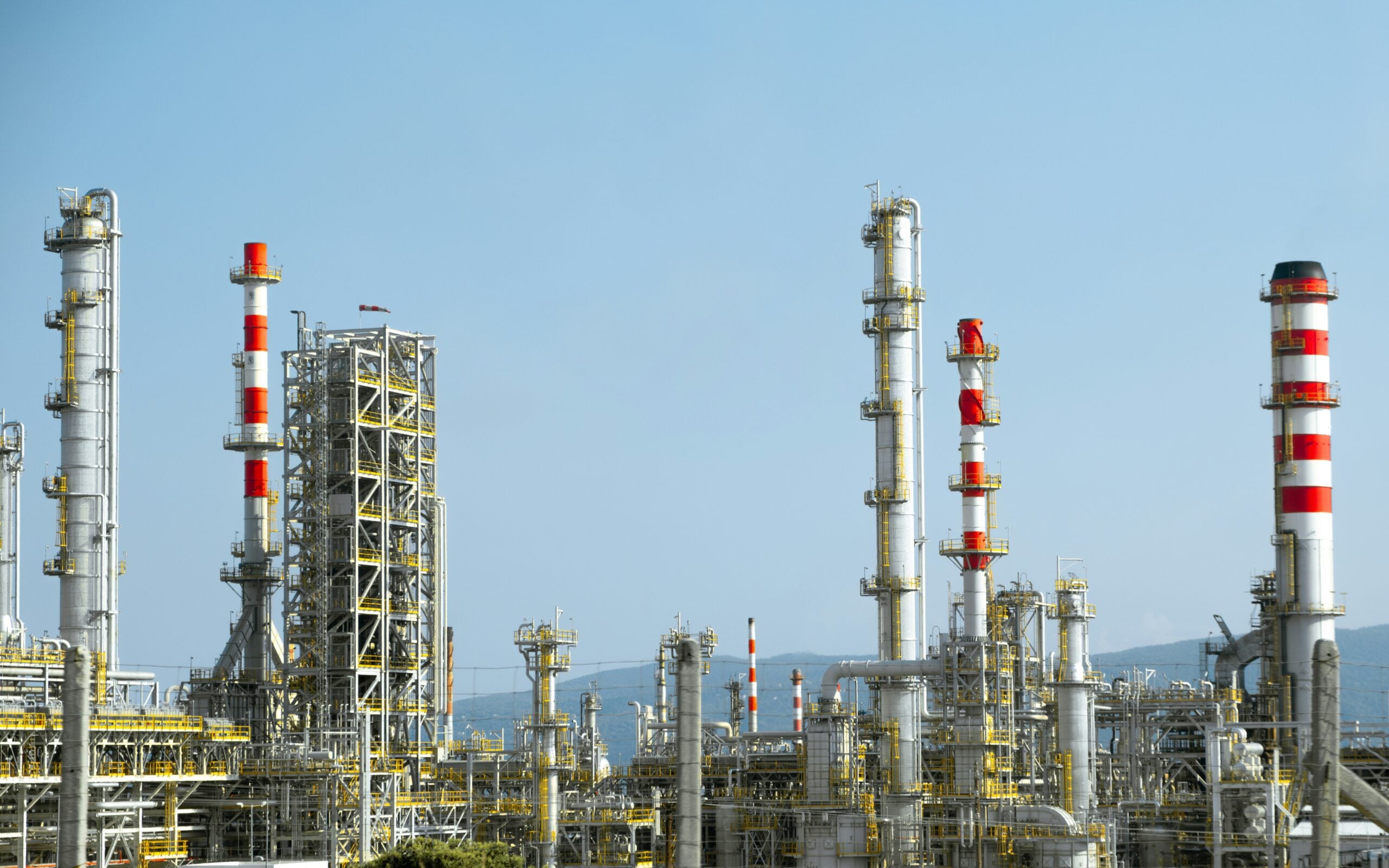 Industrial refinery complex with multiple tall distillation towers, pipes, and metal structures against a clear blue sky, with mountains faintly visible in the background.