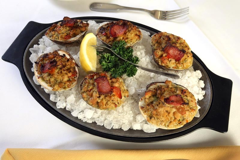 Six stuffed clams topped with bacon sit on a bed of crushed ice on a black platter, garnished with a lemon wedge and fresh parsley, with a fork and napkin nearby.