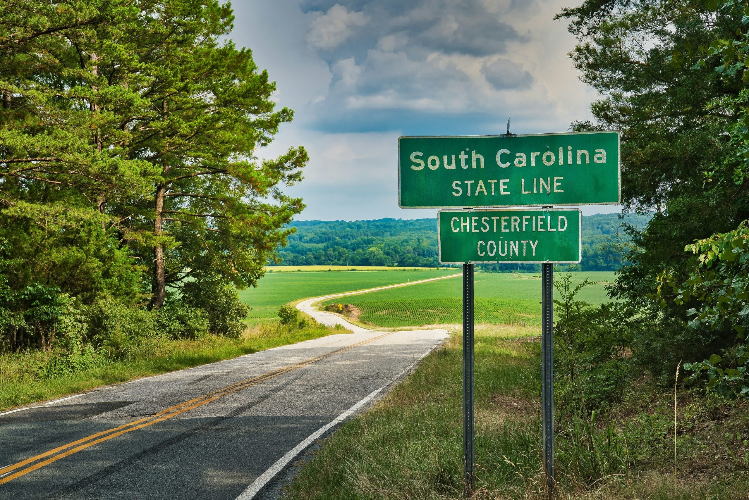 A rural road curves past a green “South Carolina State Line, Chesterfield County” sign, bordered by trees and fields under a partly cloudy sky.