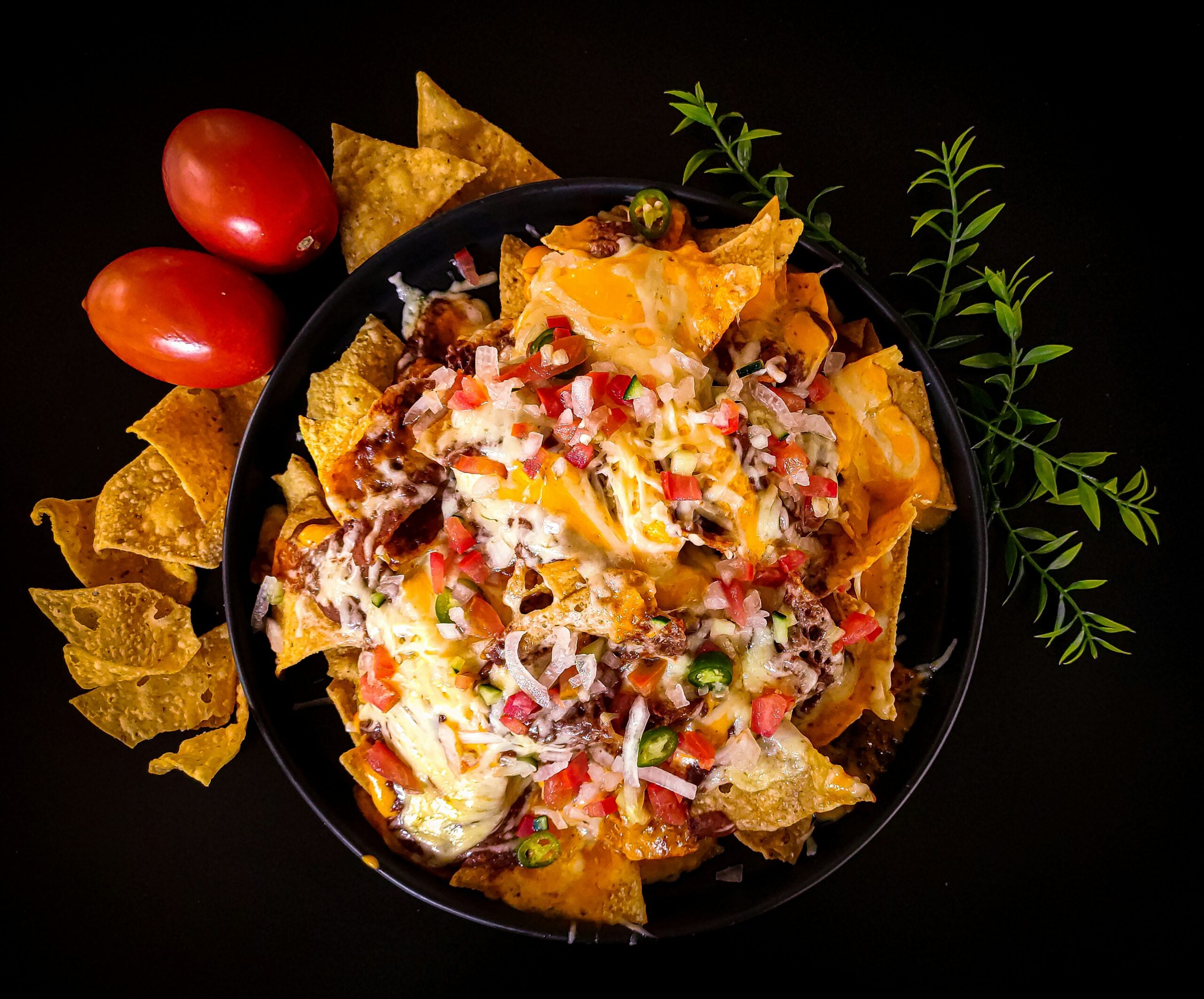 A bowl of loaded nachos topped with melted cheese, diced tomatoes, onions, and jalapeños, surrounded by tortilla chips, two whole tomatoes, and a sprig of greenery, all set against a black background.