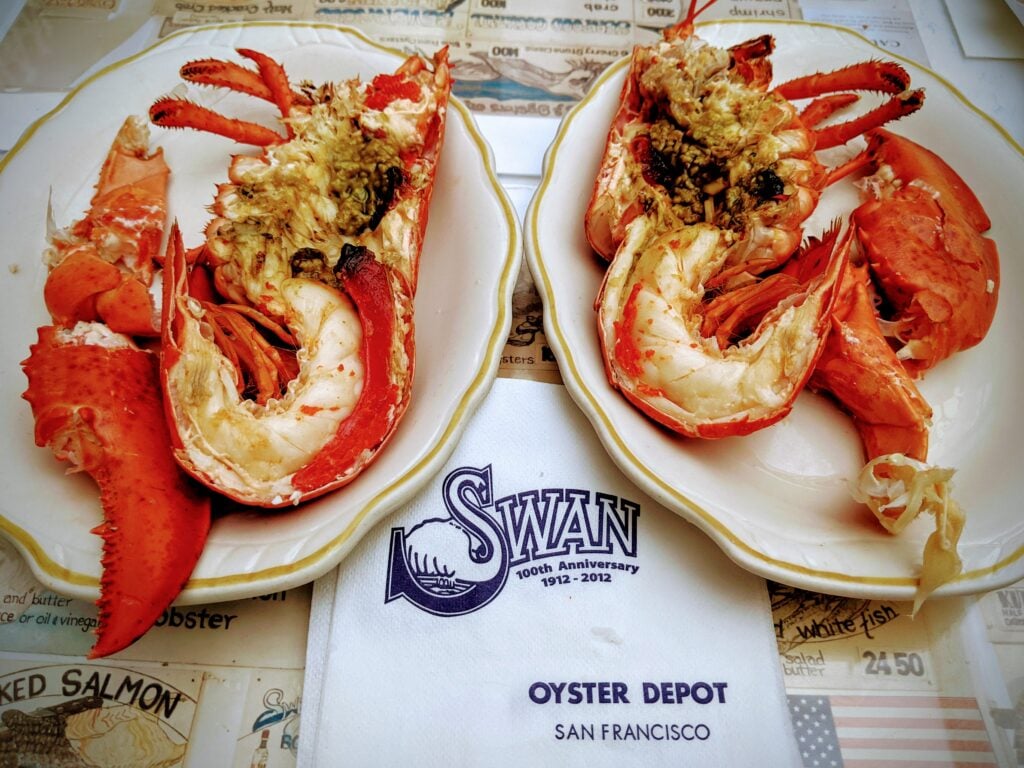 Two plates each holding a split lobster with visible claws and tails, served on a placemat and napkin labeled “Swan Oyster Depot, San Francisco.” The lobsters are garnished with herbs and seasoning.
