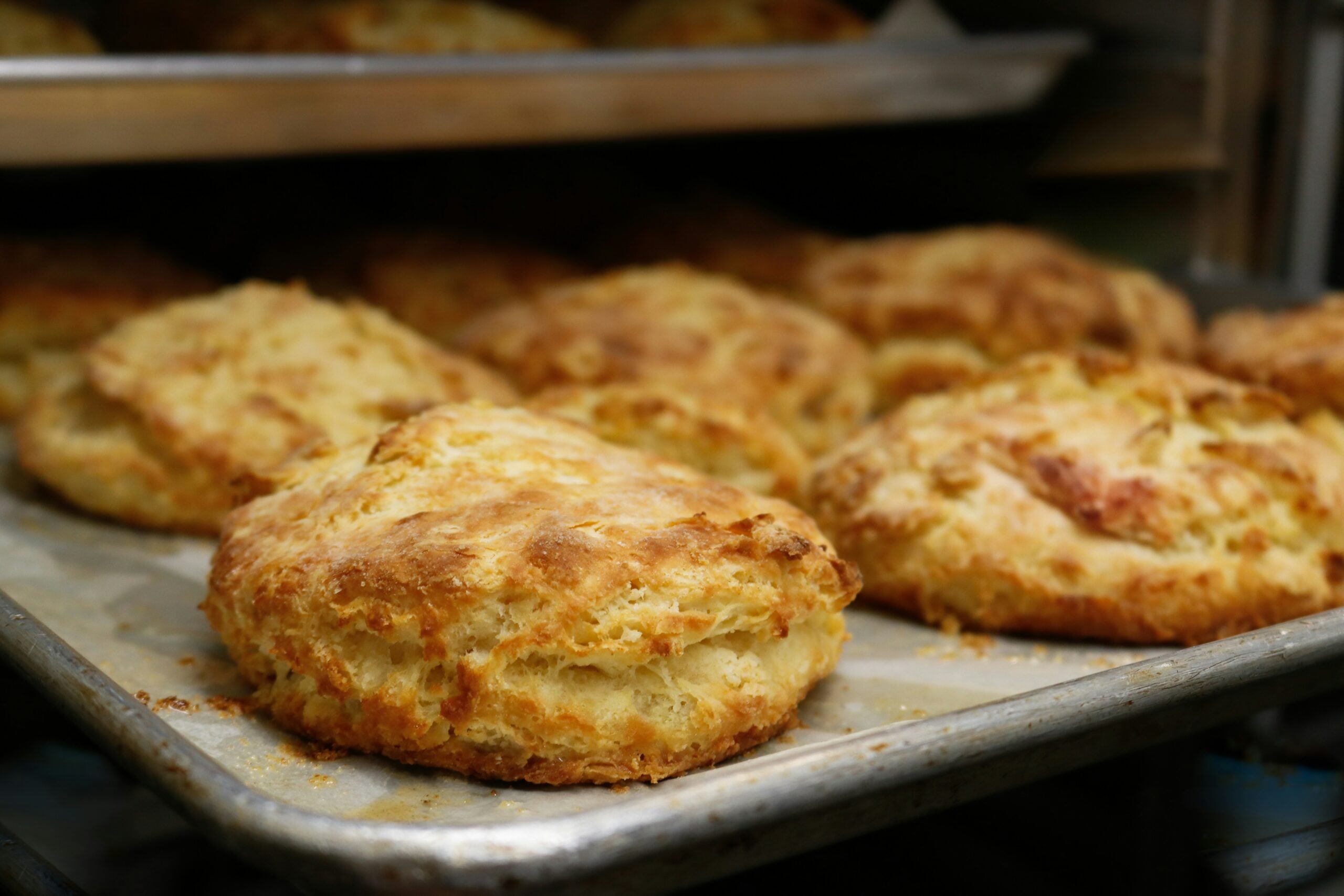 Golden brown biscuits cooling on a metal baking tray, with more biscuits visible in the background. The biscuits have a flaky, textured surface and appear freshly baked.