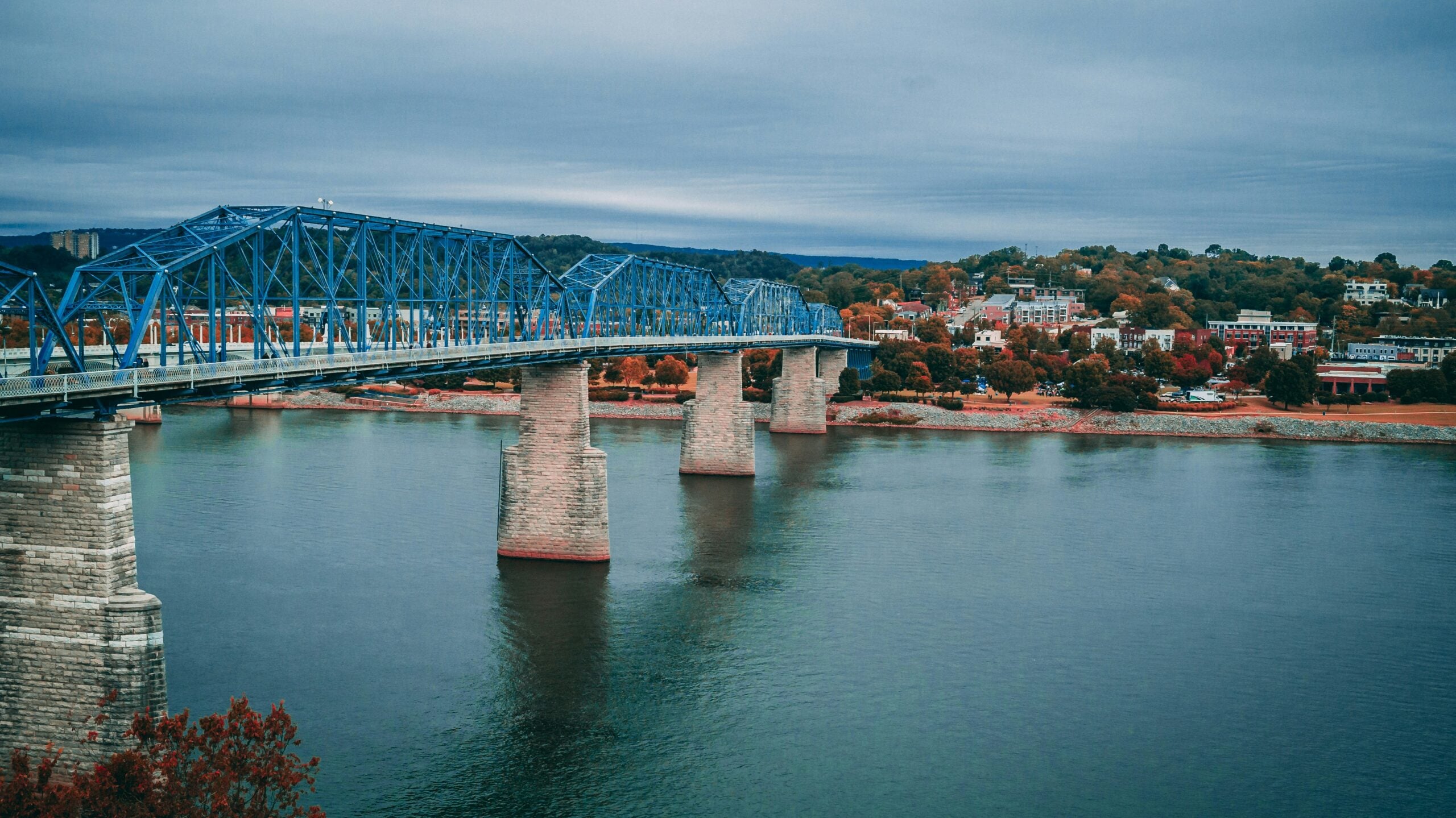 A blue truss bridge spans across a wide river, connecting two banks. On one side, a town with autumn-colored trees and buildings can be seen under a cloudy sky.