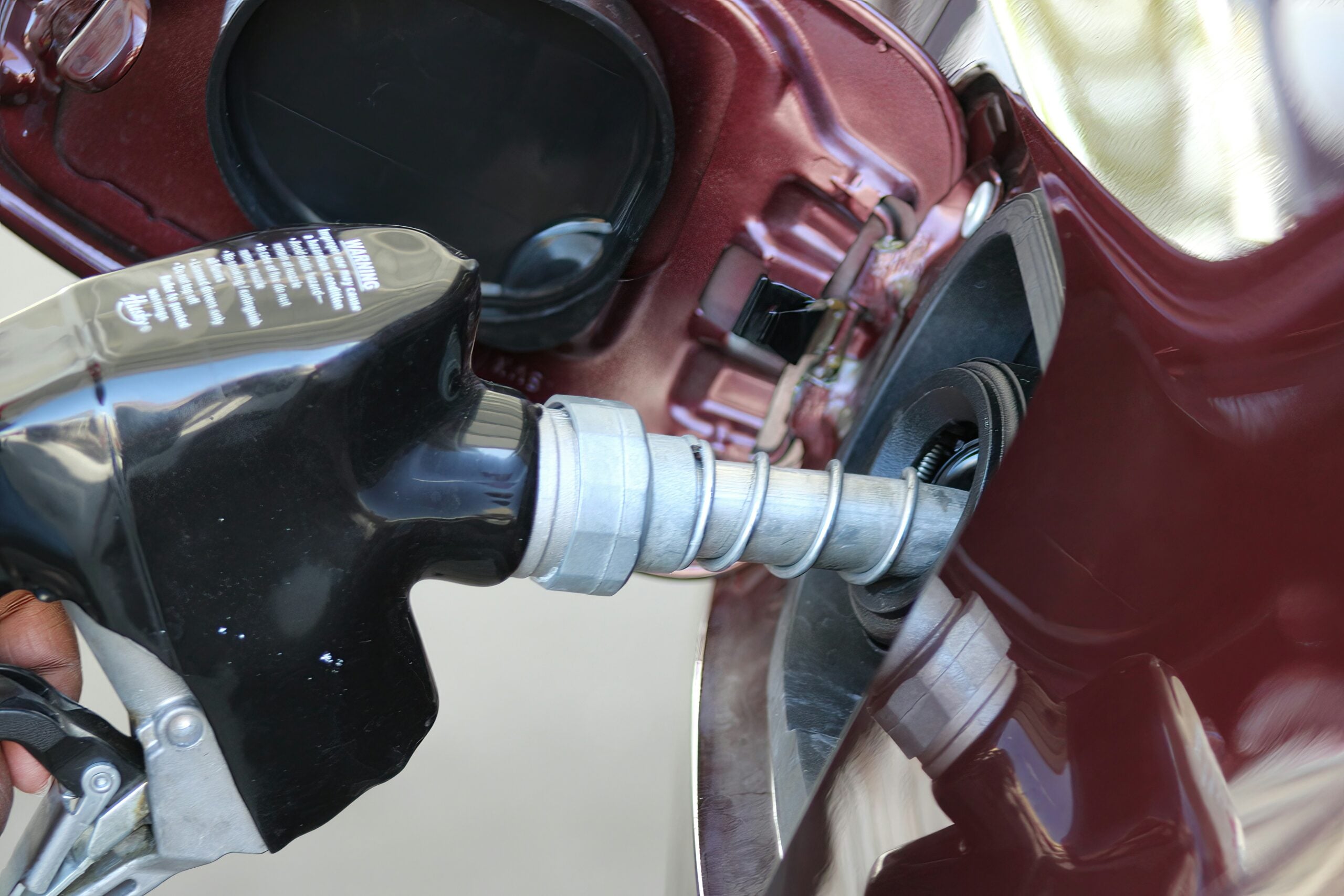A close-up of a fuel nozzle inserted into the gas tank of a maroon vehicle, with the fuel cap open, while refueling at a gas station.
