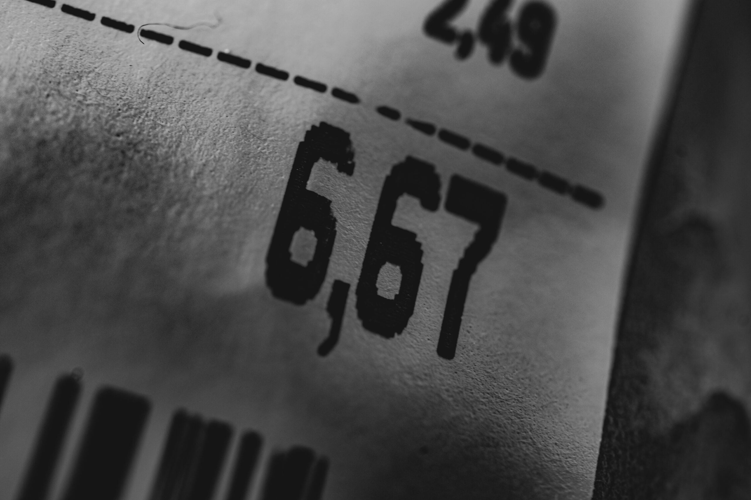Close-up of a printed price tag showing the numbers "6,67" in bold black text, with part of a barcode and another partial price visible above on a textured, white surface.
