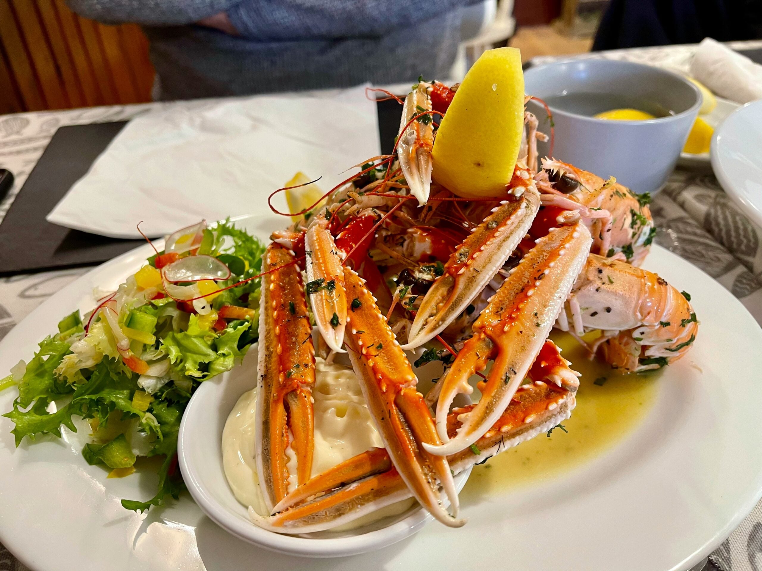 A plate of cooked langoustines garnished with herbs and a lemon wedge, served with salad and a side of mayonnaise. A person with folded arms is seated in the background.