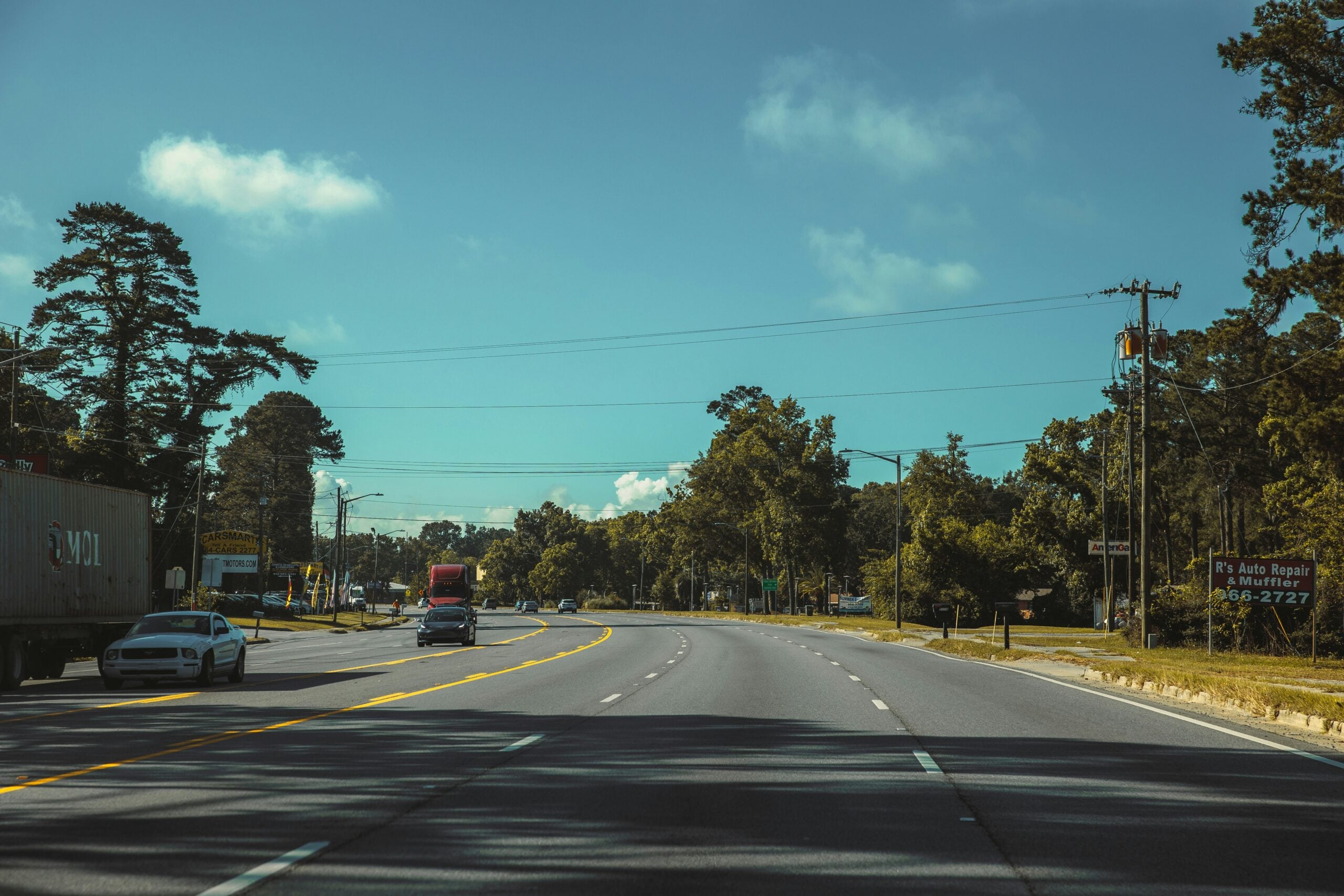 A wide, multi-lane road curves gently to the right, bordered by trees and utility poles. Cars and a truck drive under a blue sky with scattered clouds on a sunny day.