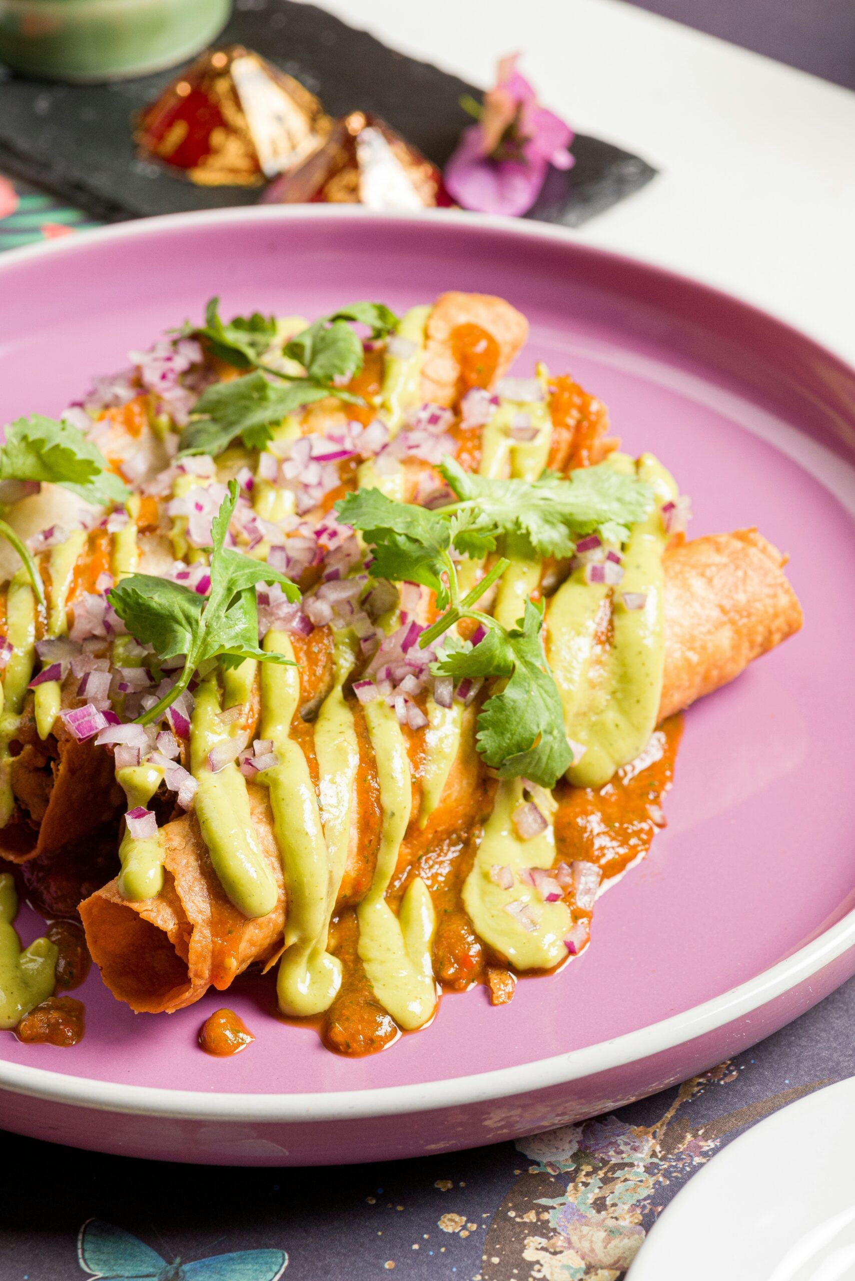 Three rolled, crispy taquitos topped with green avocado sauce, chopped red onion, fresh cilantro, and drizzled with salsa, served on a pink plate. In the background, dessert pieces are visible on a black tray.