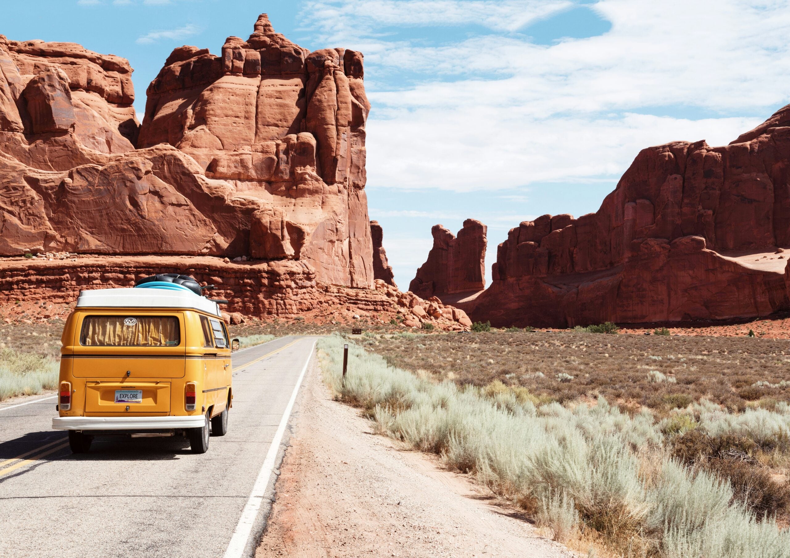 A yellow van with a blue kayak on top drives down a road through a desert landscape with large red rock formations under a partly cloudy blue sky.