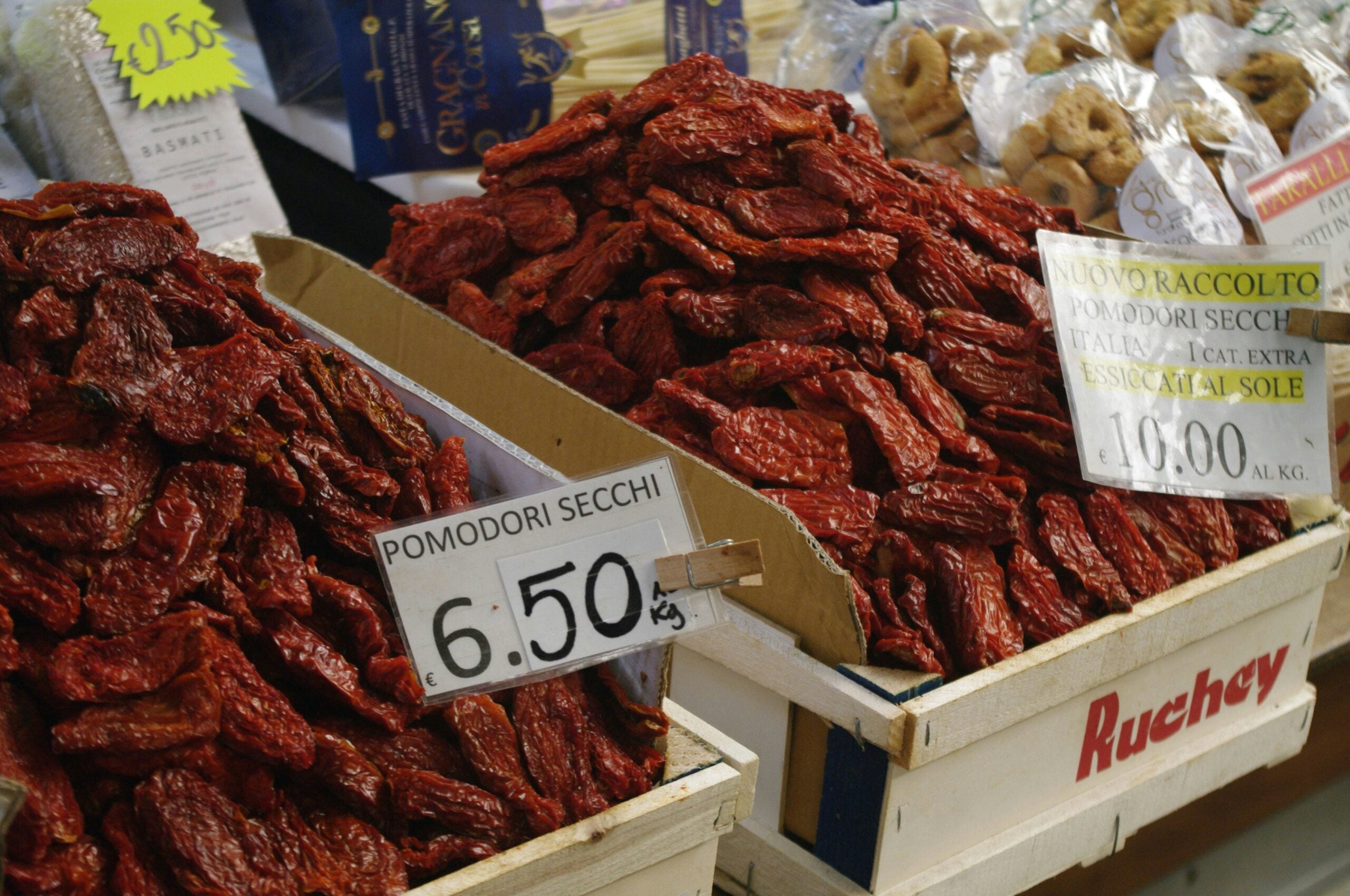 Piles of sun-dried tomatoes are displayed in wooden crates at a market, with handwritten signs showing prices per kilogram in euros. Bags of other dried foods are visible in the background.
