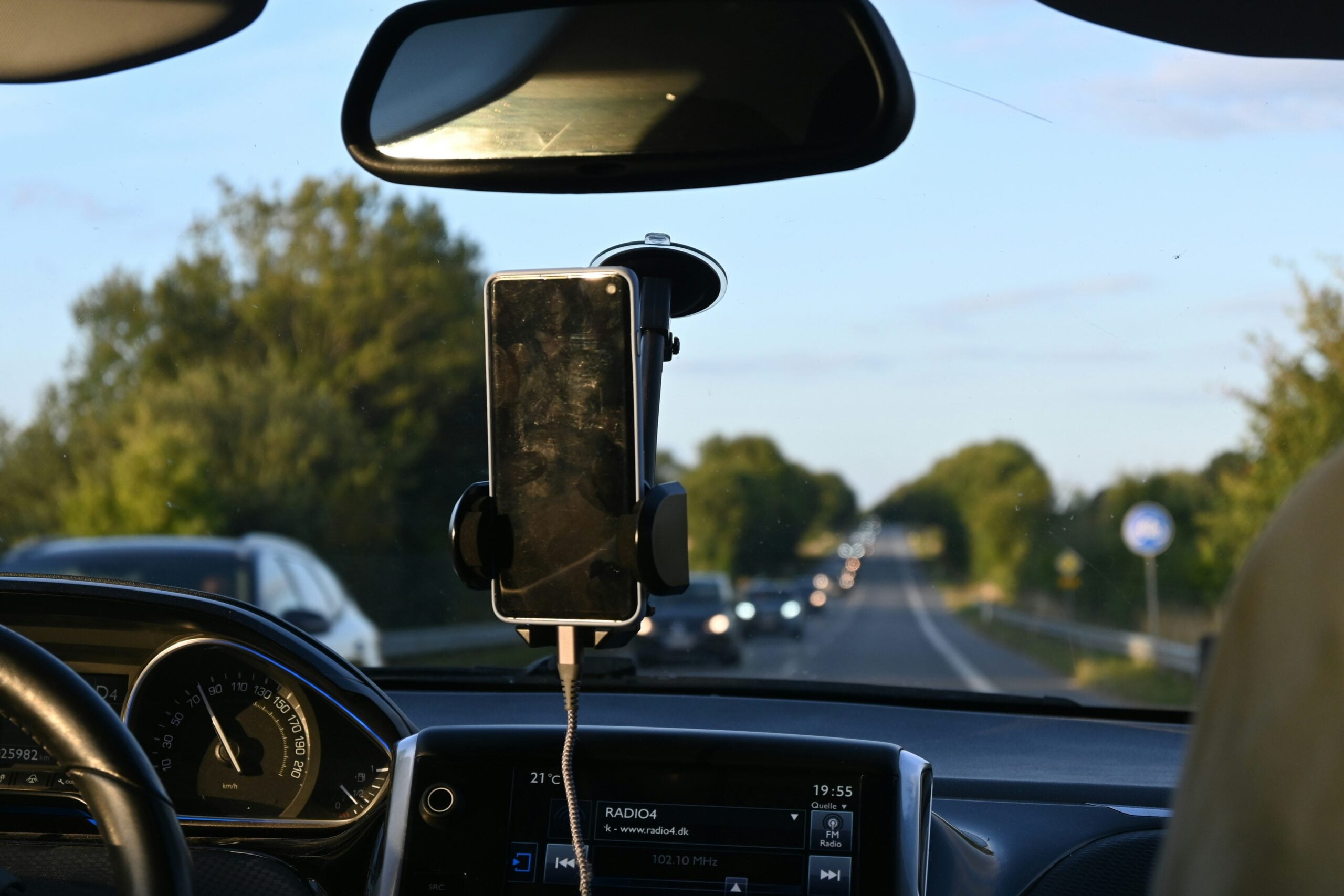 A smartphone is mounted on a car dashboard, displaying a dark screen. The car is traveling on a road with trees on both sides, and other vehicles are visible ahead through the windshield.