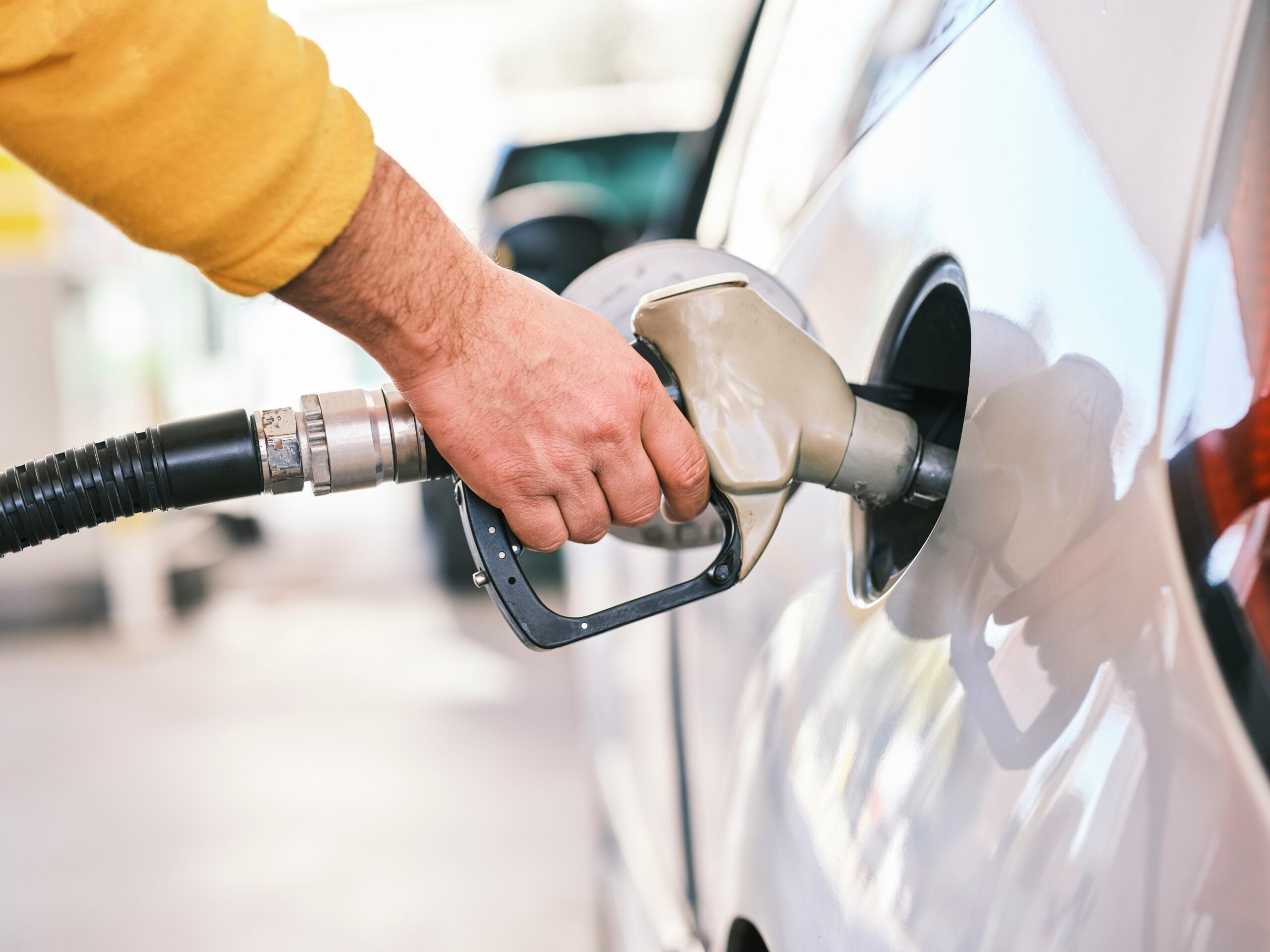 A person wearing a yellow sleeve is holding a fuel nozzle, filling up a silver car at a gas station. The focus is on the hand, nozzle, and car's fuel tank.
