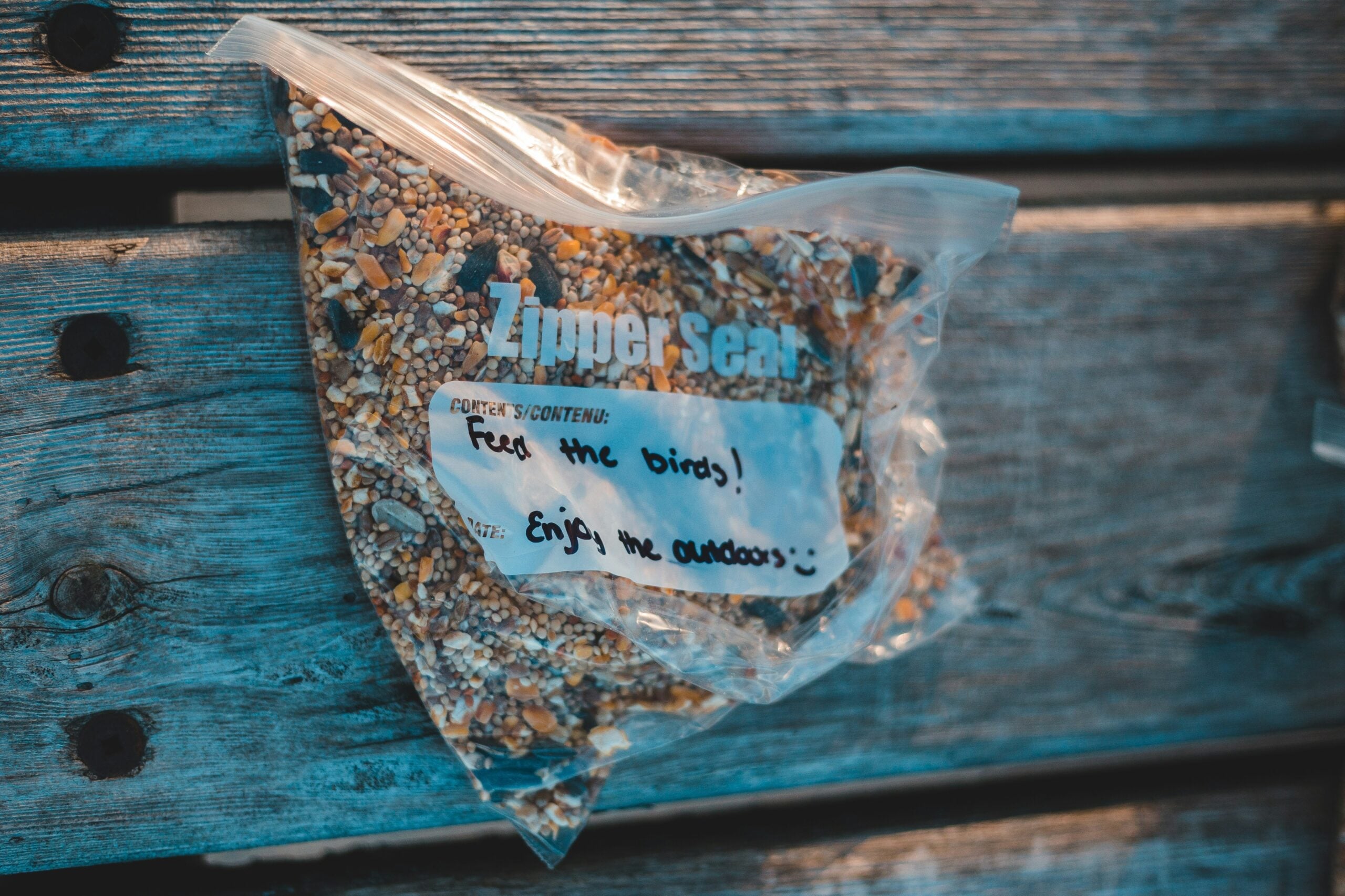 A plastic zipper bag filled with birdseed rests on a wooden surface. A label on the bag reads, "Feed the birds! Enjoy the outdoors :)" written in marker.