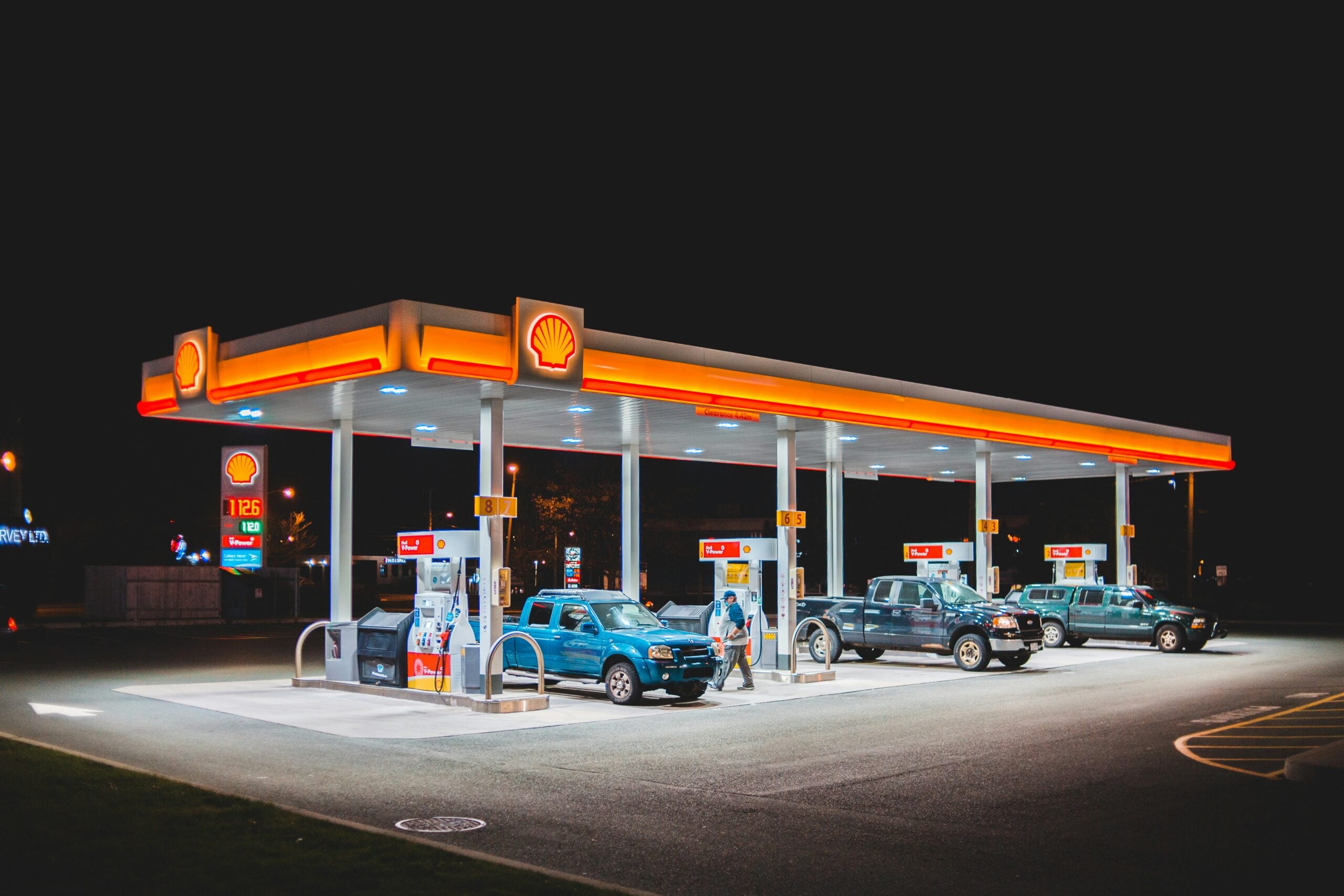 A brightly lit Shell gas station at night with several cars and trucks parked at the pumps, and people refueling their vehicles under the illuminated canopy.