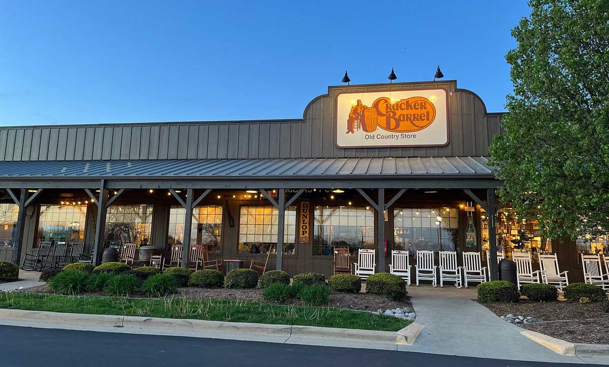 Cracker Barrel Old Country Store exterior at sunset, with a large porch, rocking chairs, string lights, and a prominent sign above the entrance. Guests chat about gas prices as they stroll past shrubs and trees lining the walkway in front. Long-running restaurant chains.