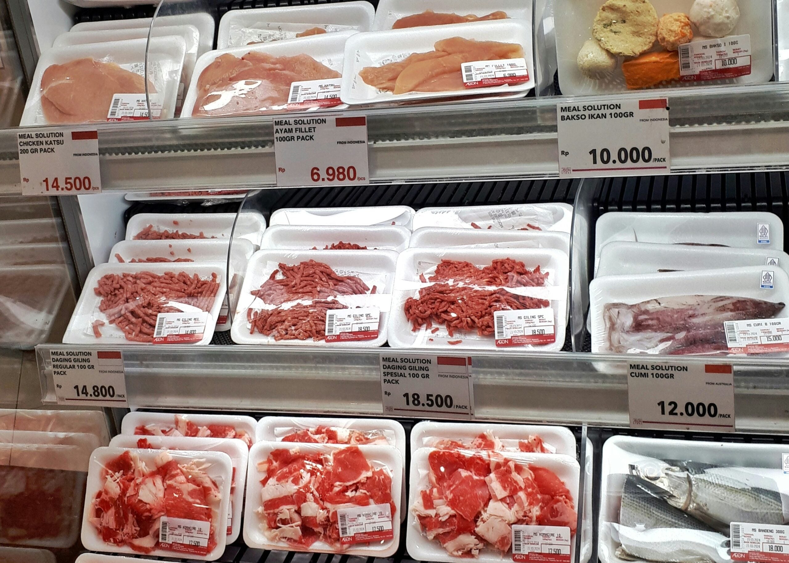 A supermarket meat display with packaged cuts of beef, chicken, and fish in white trays with price labels. Prices range from 6,980 to 18,500, and some trays are labeled “MEAL SOLUTION.”