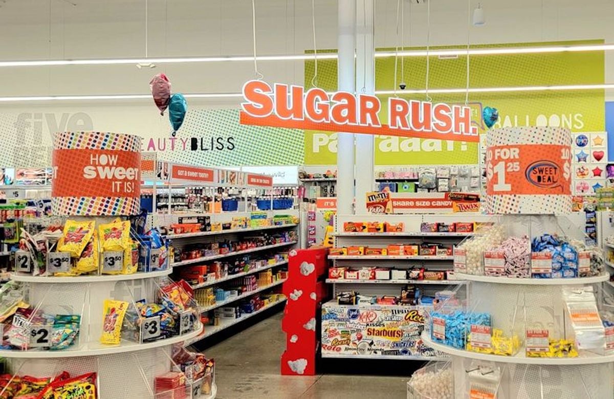 A brightly lit store aisle features shelves stocked with candy and snacks. For a first time at Five Below, the large “Sugar Rush” sign and colorful displays with deals like “2 for $1” make the atmosphere extra inviting.