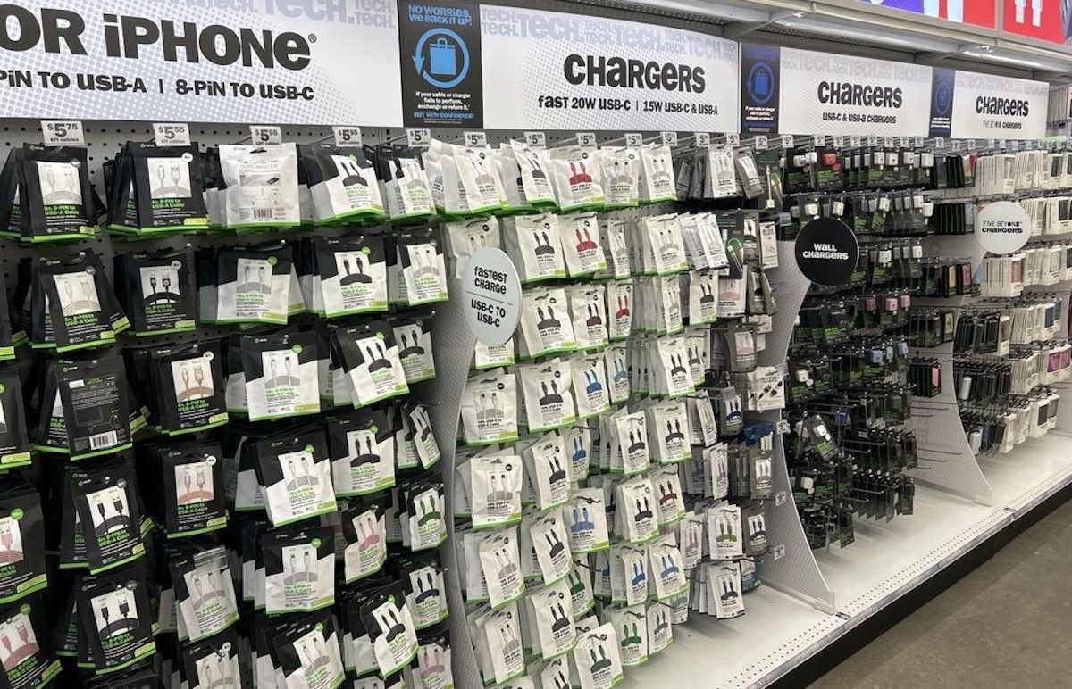 A store shelf at Five Below, stocked with various phone chargers, USB cables, and charging adapters in white and black packaging, organized by type and labeled for iPhone and other devices. Signs above indicate different charger options.