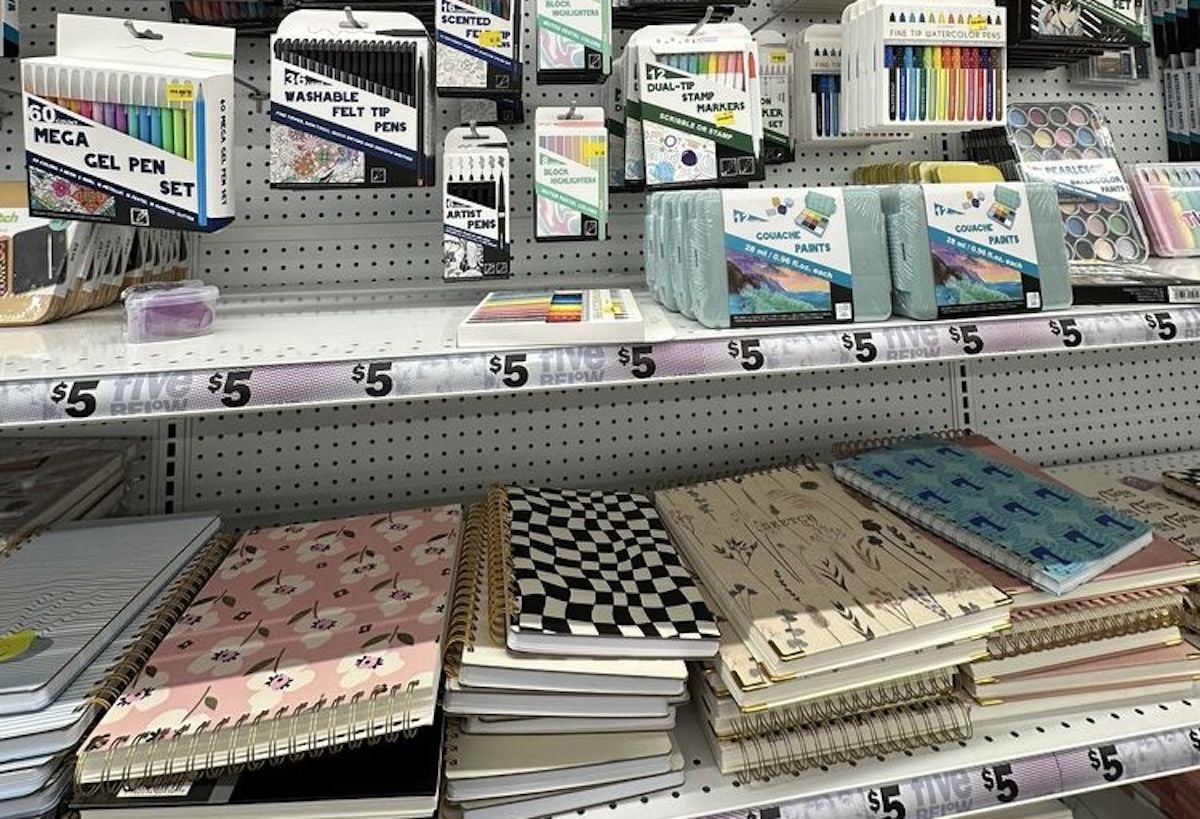 Shelves in a store display various stationery items, perfect for your first time at Five Below—choose from notebooks with unique covers, packs of pens and markers, and art supplies. Each item is just $5.